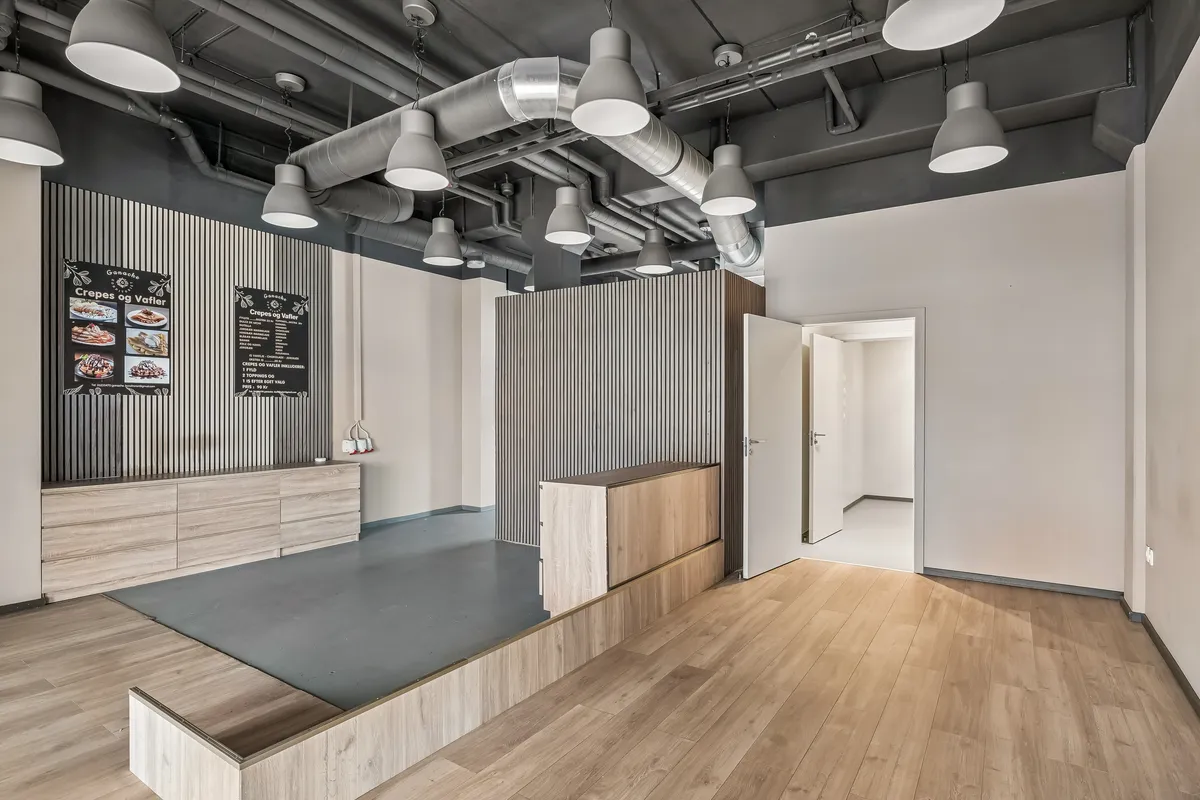 Restaurant med moderne interiør på Toftegårds Allé 5A. Lejemål med lyse trægulve og lysekroner. Væg med menukort og åben dør til køkken.