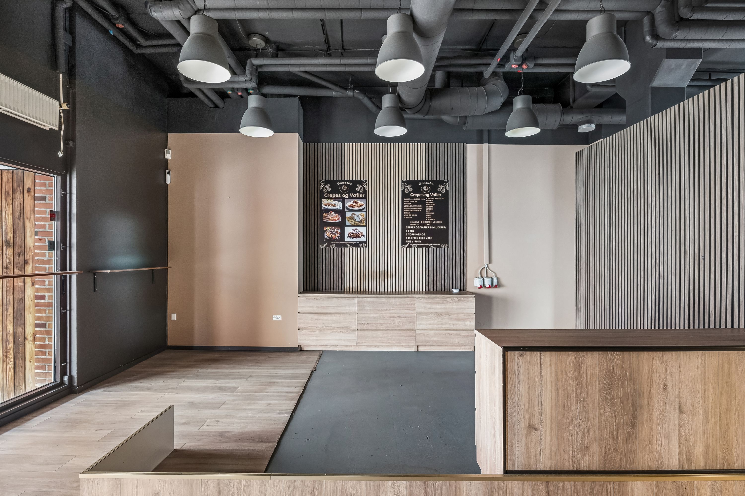 Moderne og rå indretning i restauranten med hængelampe og trægulv samt menutavle på væggen og en udgang til en gård med røde mursten og glasdør