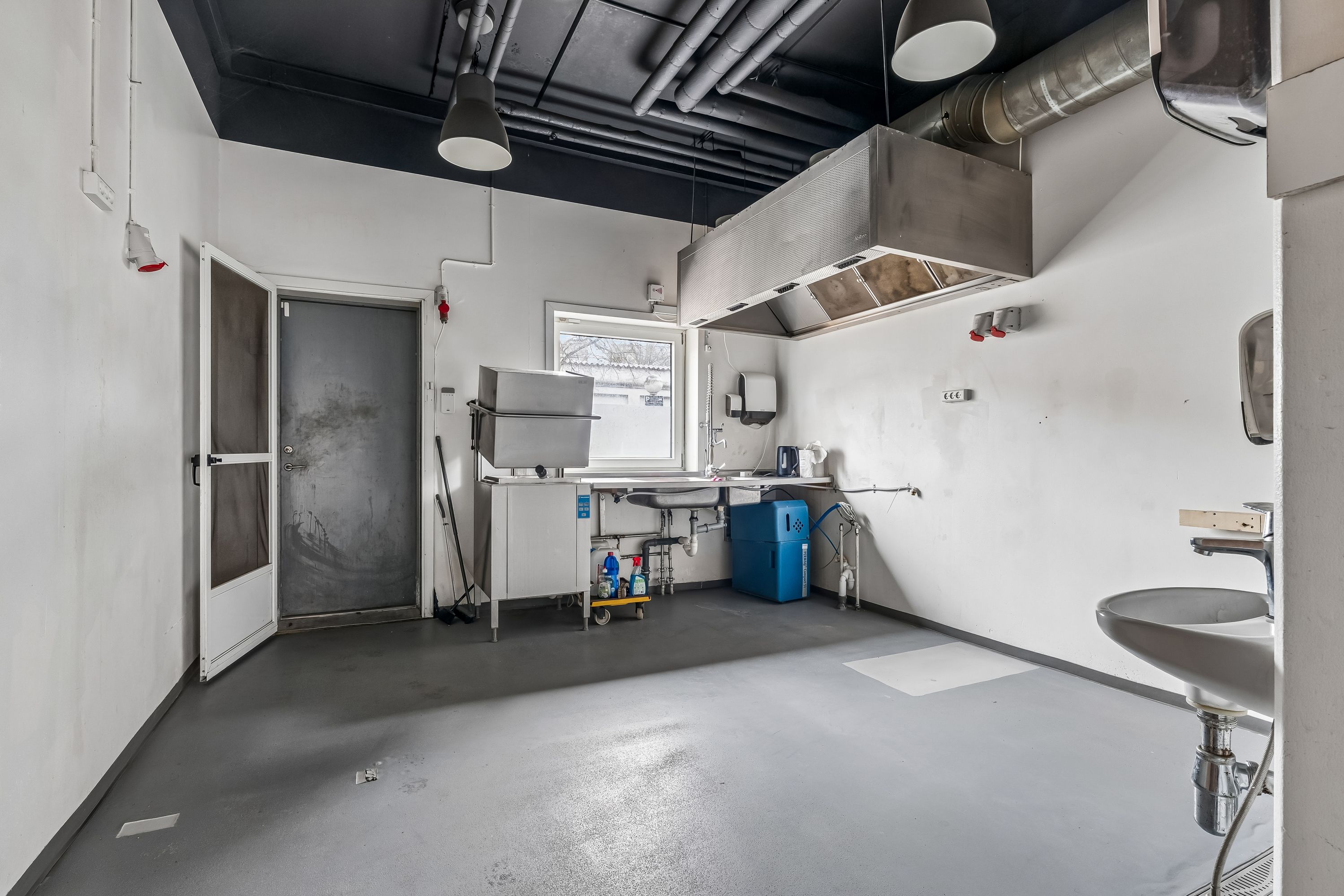 Rådkøkken med højt loft, lysekroner og hængende metalventilator. Der er en strygejern og en opvaskemaskine. Der er flere døre, og der er en gulvskrubbe.