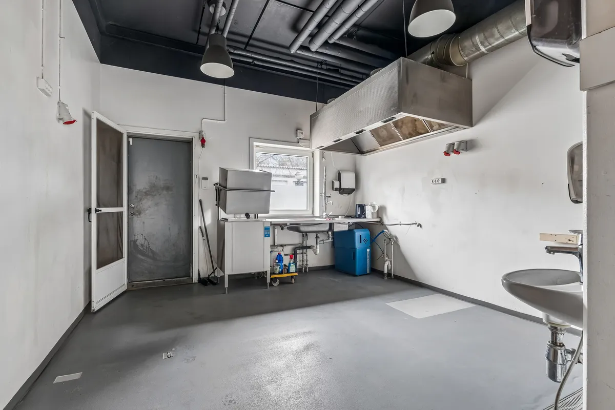 Rådkøkken med højt loft, lysekroner og hængende metalventilator. Der er en strygejern og en opvaskemaskine. Der er flere døre, og der er en gulvskrubbe.