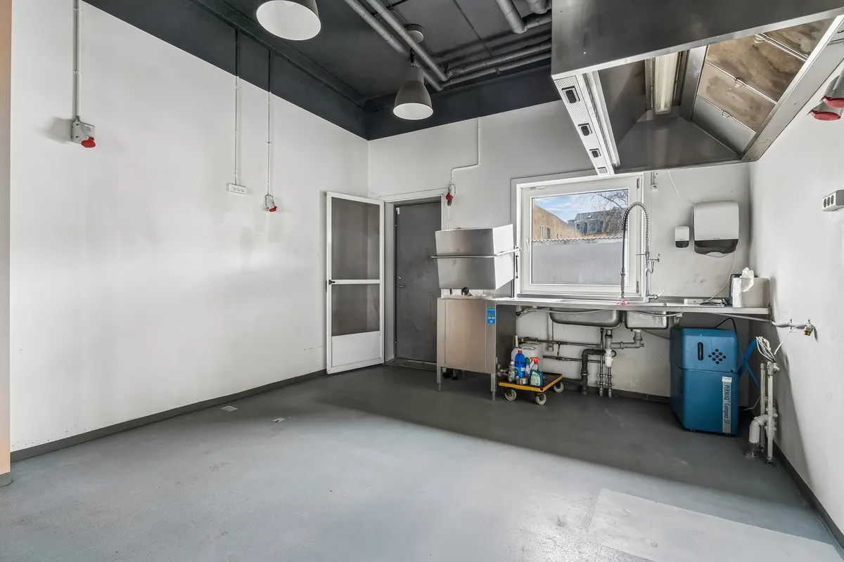 Lejlighed med rå og moderne indretning. Der er et køleskab, 2 senge, et vindue og et køkken med 2 vandhaner og en skraldkurv.