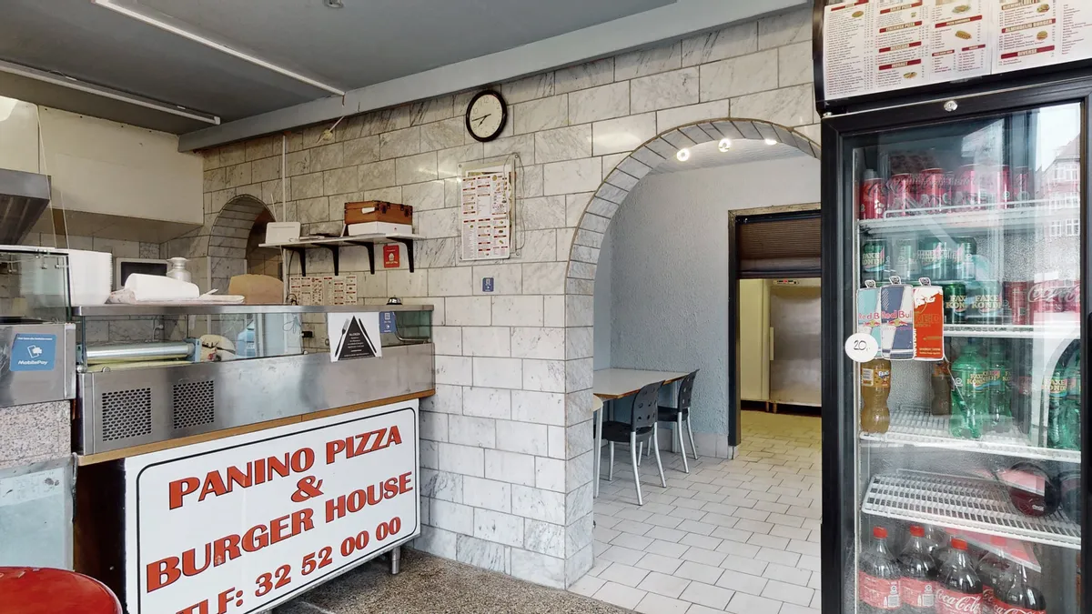 En hjørnerestaurant med skrivet Panino Pizza & Burger House. Man kan se en køkkenbord, en køleskab med sodavand og en klokke på væggen. Der er to sæde og en bord i en sammenhængen.