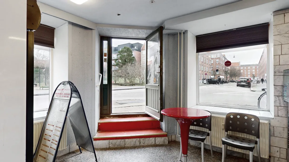 Foto af en erhvervsandel med åbne døre og røde trin. En menu og en rødt rundt bord med to stole er placeret inde i lokalet. Et søjleskilt er synligt bag dørene.