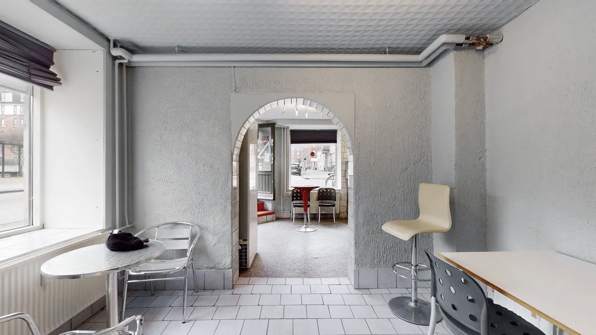 Lukket forretningslokal med moderne møbler, højt loft og lyse gulvfliser. Bagved er en åben bue og en dør, der fører ud til en gadekøkken.
