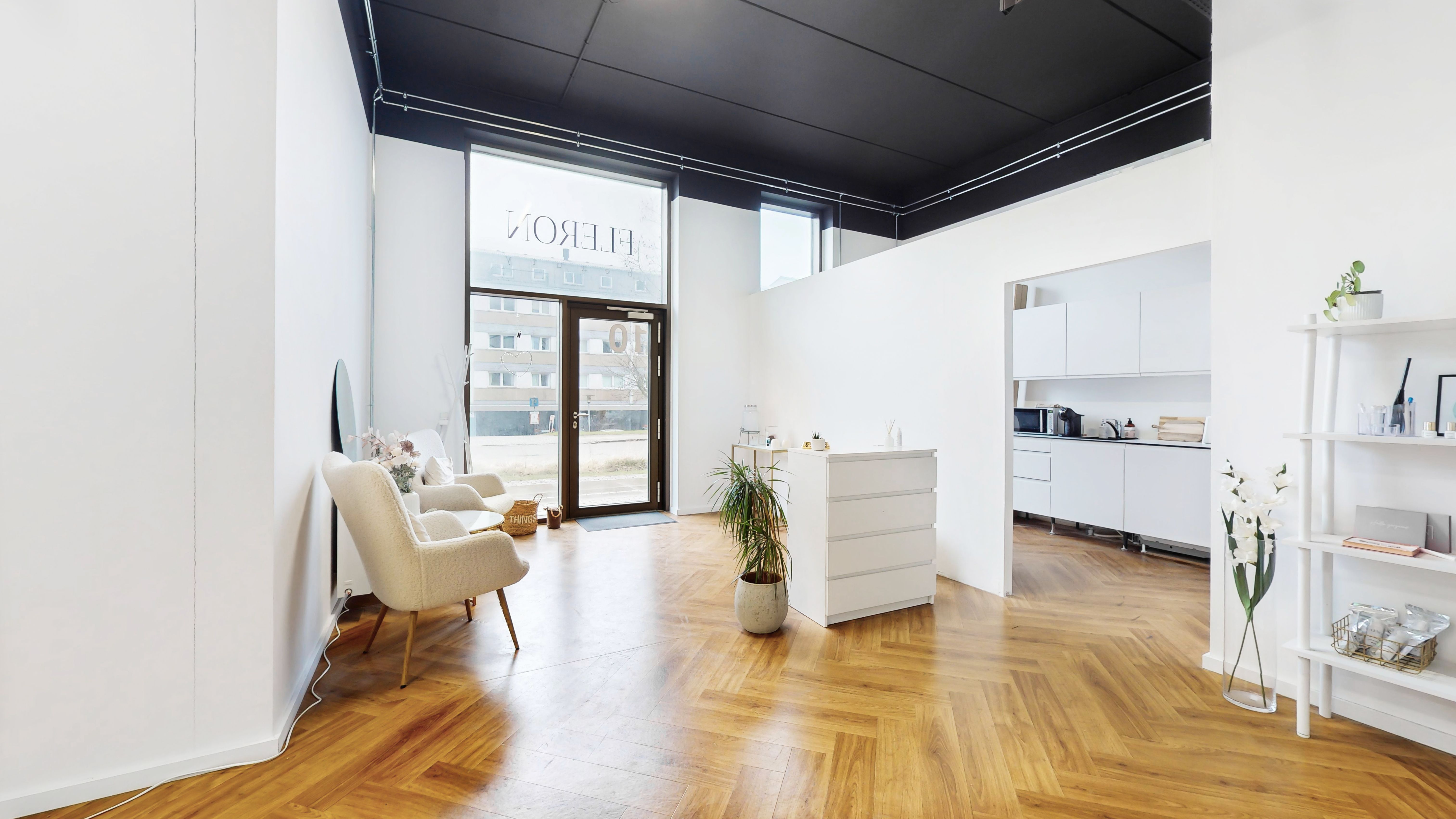 Kontor i stueplan med hvide vægge, trægulv og lyse vinduer. Væggen er præget med et navn. Der er en hvid sofa med pude og en hvid højbord med plante og vase med blomster