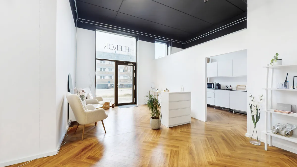 Kontor i stueplan med hvide vægge, trægulv og lyse vinduer. Væggen er præget med et navn. Der er en hvid sofa med pude og en hvid højbord med plante og vase med blomster