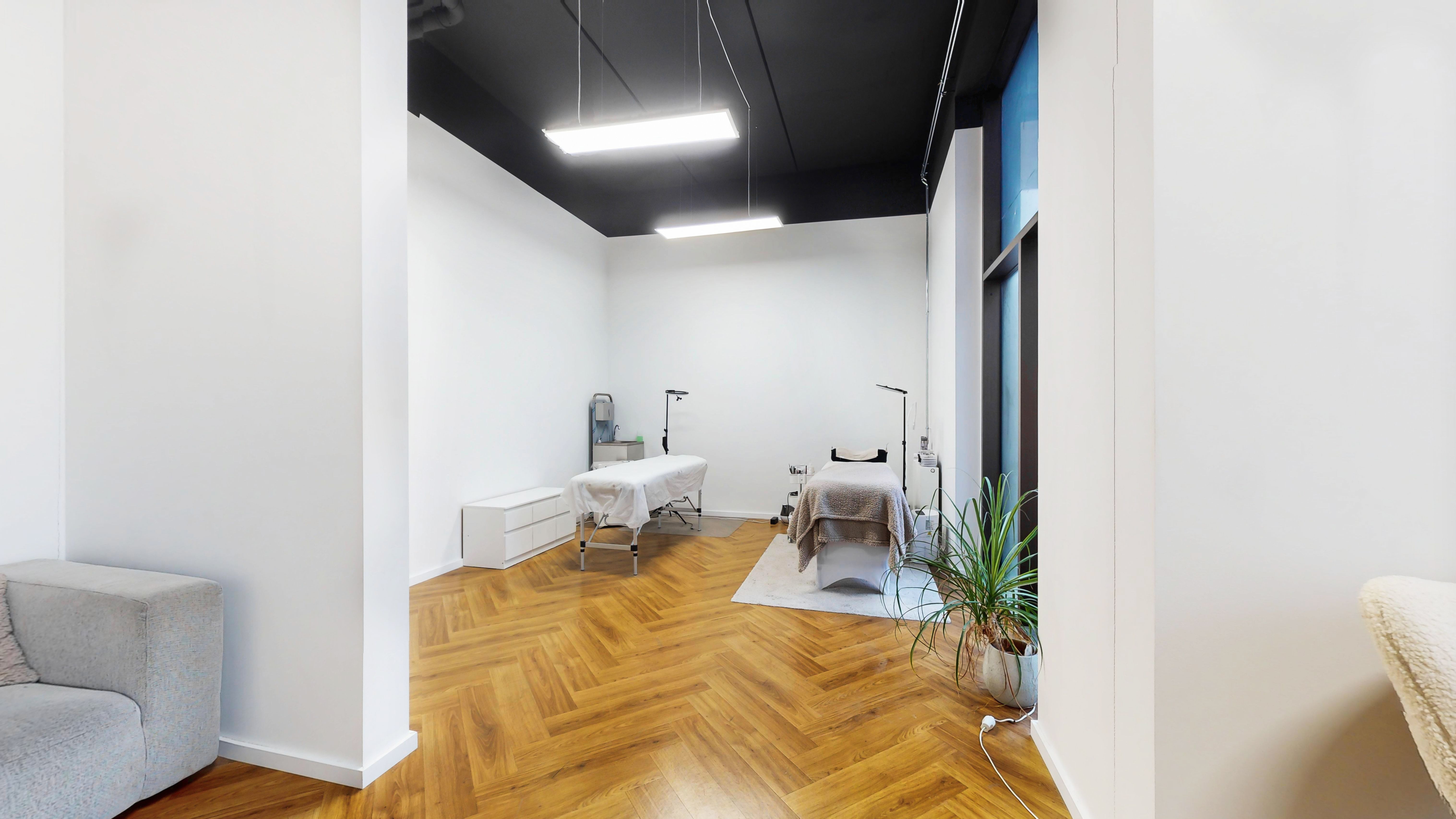 En rummelig og veludstyret massagesalon med to massageborde, en hvid sofa, hvide vægge og trægulv, samt en stor plante og et vinduesparti med udsigt til gaden