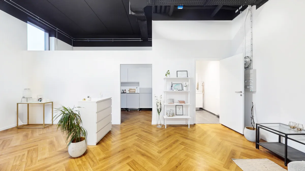 Et lille kontor med hvidt møbel og plante. Køkken med køleskab og højt højre skur. Hvid væg og gulv med vinduesåbningen. Forskellige hylder og objekter. Et enkelt gulvtæppe og et glasbord.