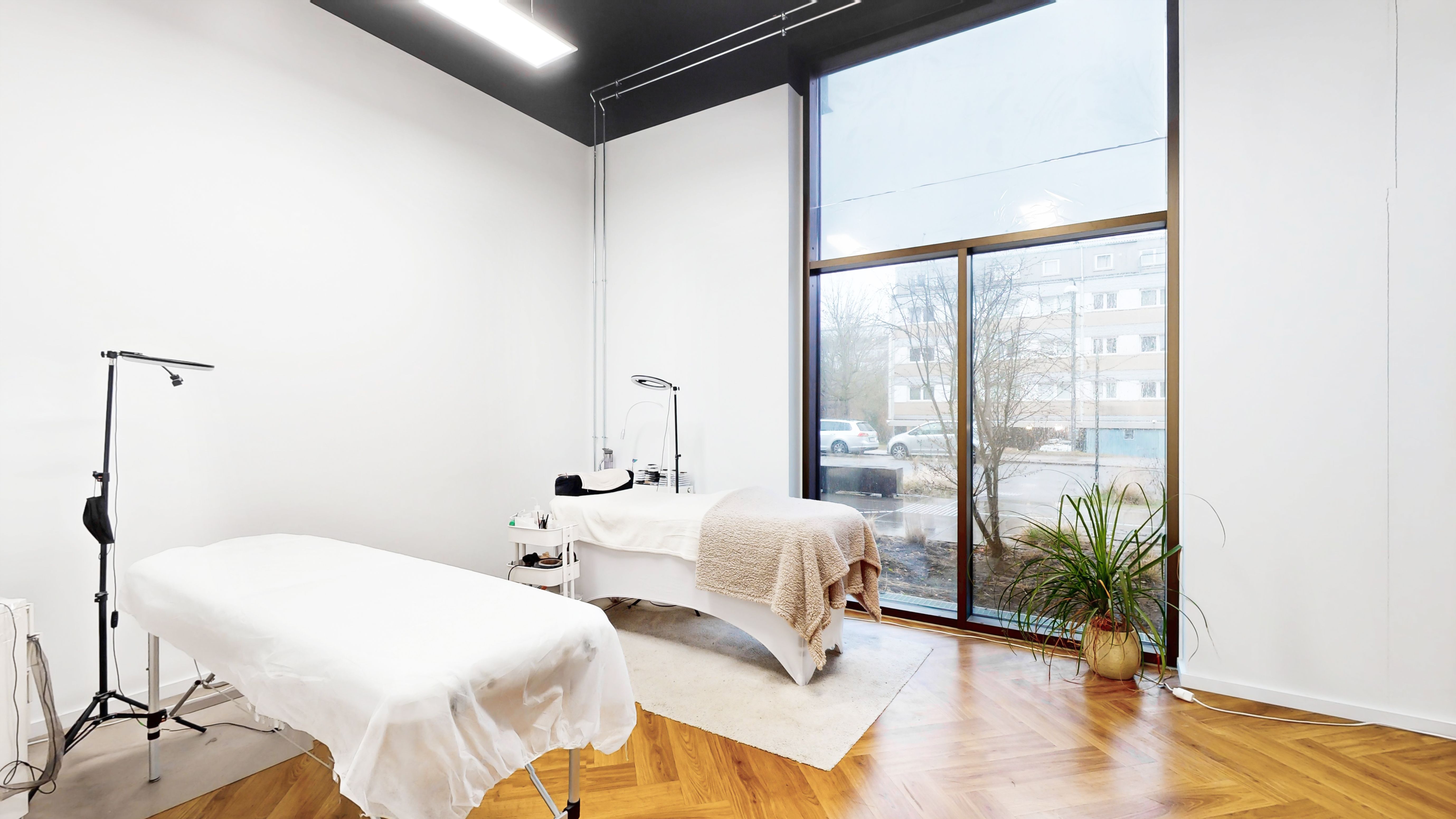 Lyst og funktionelt kontorlokum med moderne interiør og to massagebord dækket af hvide plastforslag. Plante ved vinduet og højt gulv giver en komfortabel arbejdsplads