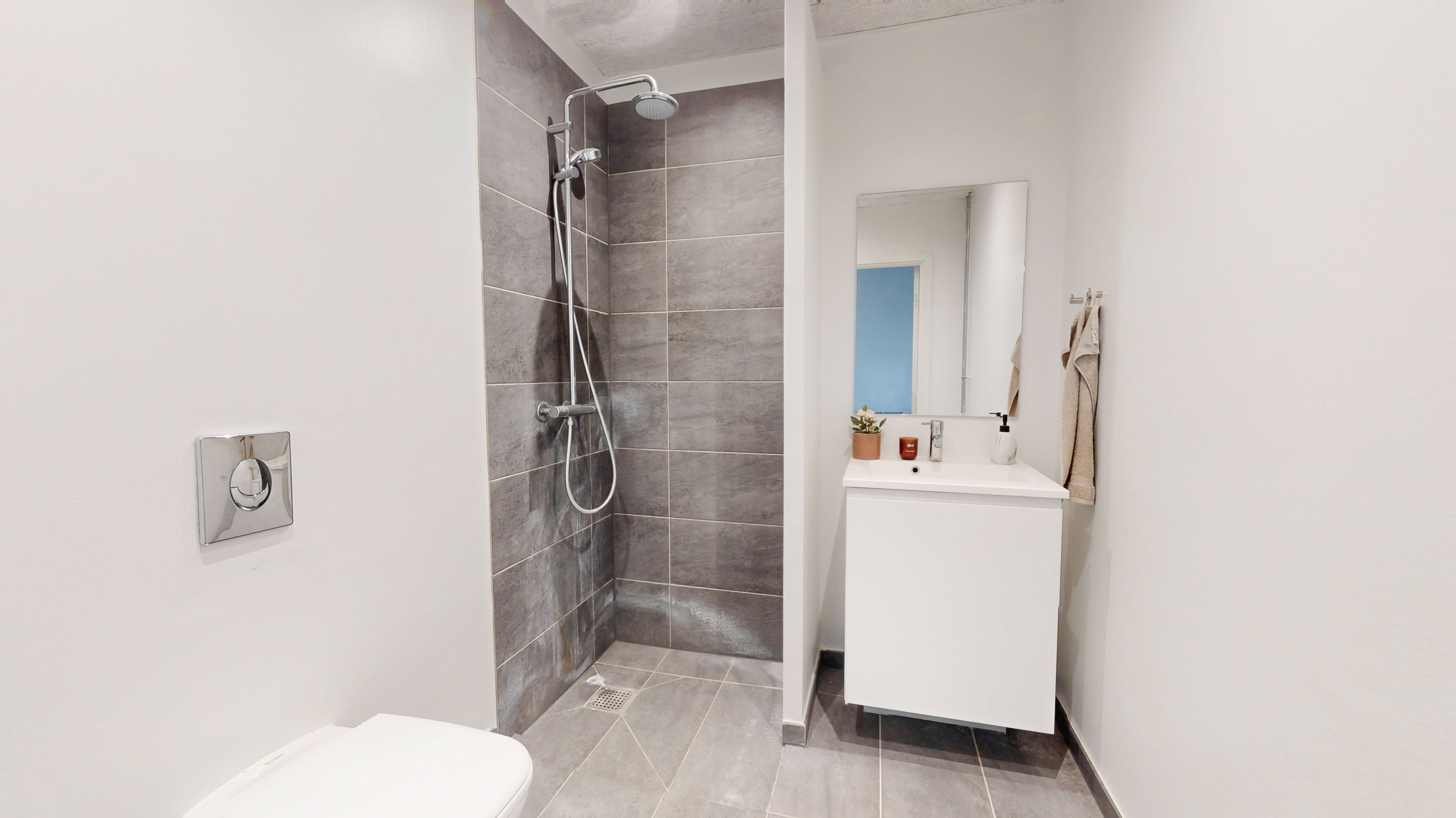 Ren og moderne badeværelse med hvid toilet, hængende spejl, hvid vask med plante og lyse grå flise og brusebad med højtrykssprøjte