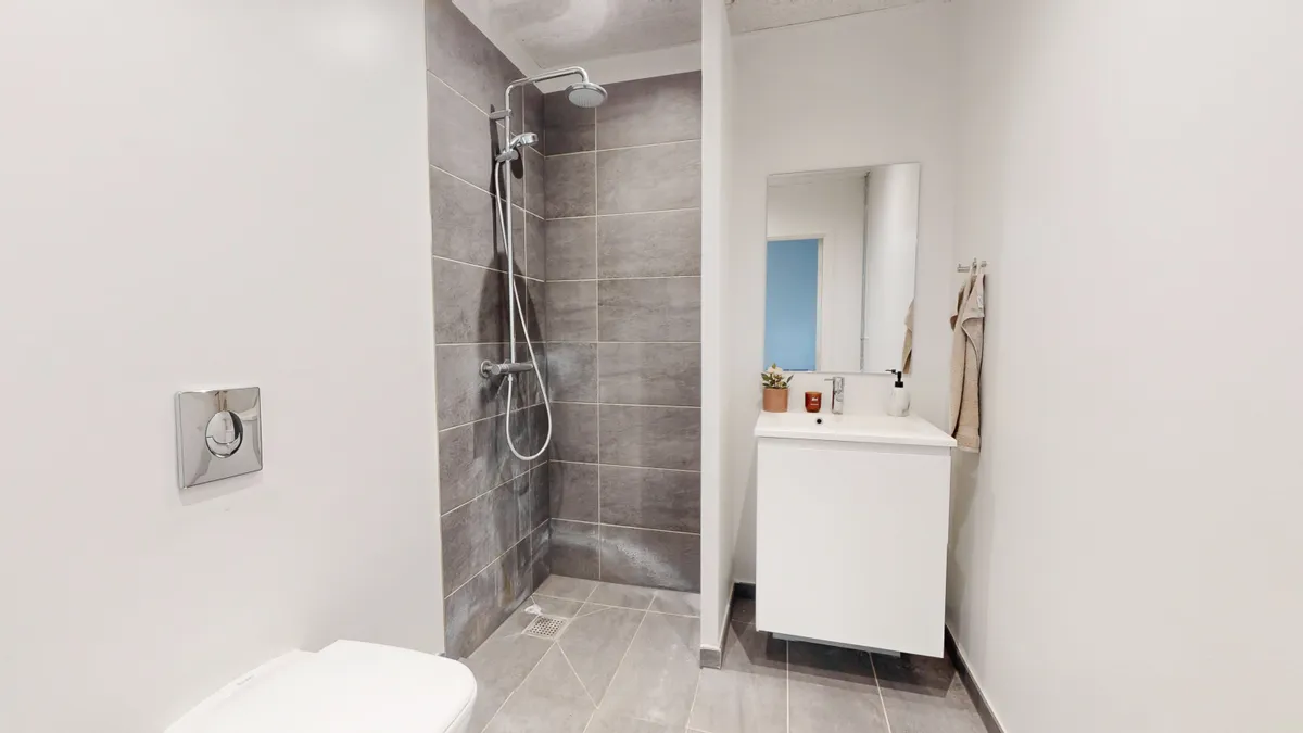 Ren og moderne badeværelse med hvid toilet, hængende spejl, hvid vask med plante og lyse grå flise og brusebad med højtrykssprøjte