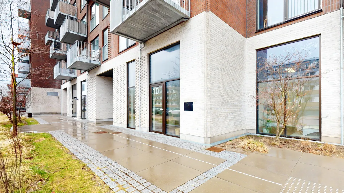 Kontorbyggeri med glasvinduer, terrasser og en vej, der fører ned ad en skråning mod en skovkant. En grøn buske og et træ er plantet på kanterne.