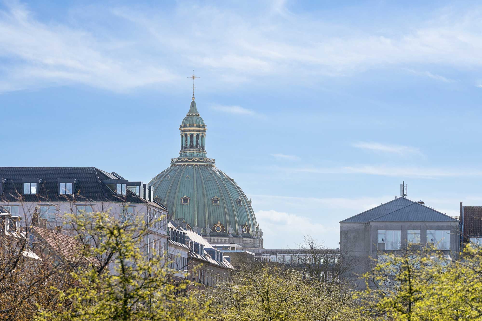 Kontorbyggeri med smukke træer og lyse vindue i en bygning med en kuppel og spire, hvor man ser en kirke i baggrunden under en lys, blå himmel med skyer