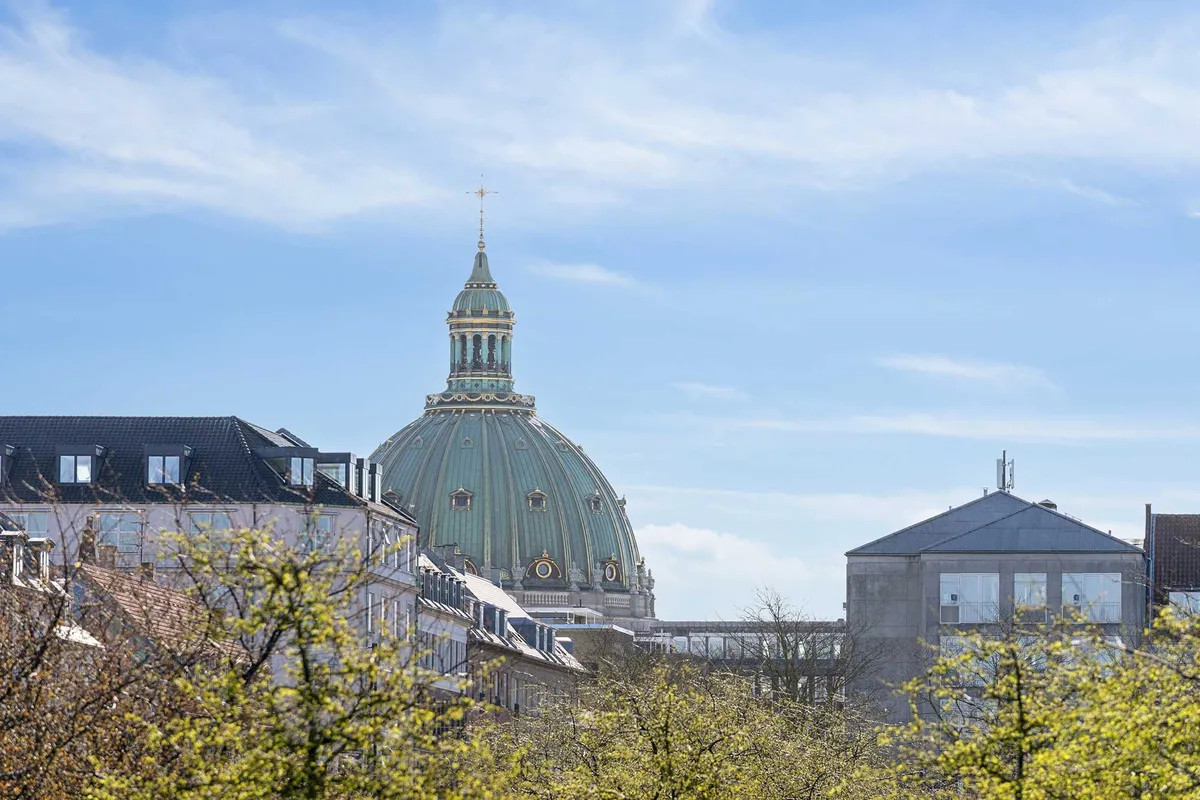 Kontorbyggeri med smukke træer og lyse vindue i en bygning med en kuppel og spire, hvor man ser en kirke i baggrunden under en lys, blå himmel med skyer