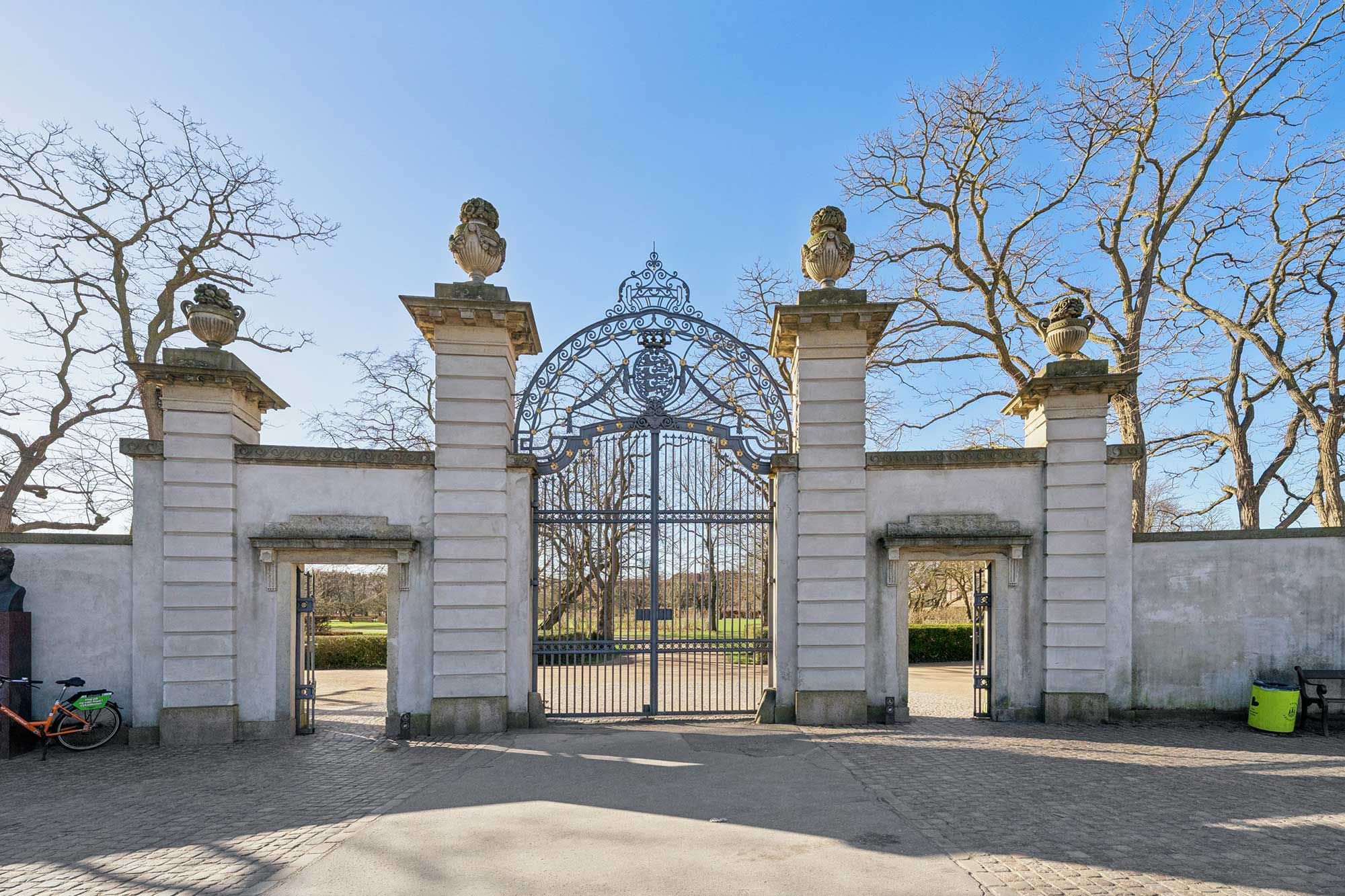 Ornate smedejernsportal med skulpturer og statuer på hjørnepiller og en statue på en sokkel med en cykel til venstre og et skraldespand til højre