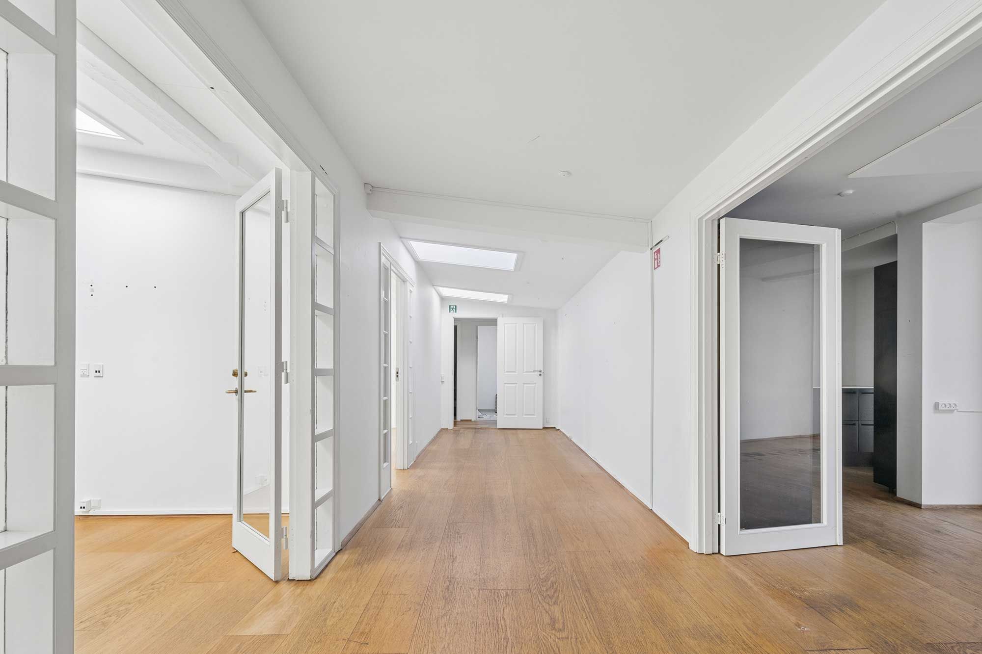 Kort og lys hallvej i et kontorlejemål med hvide vægge og lyse lofter. Trægulve og glasdøre til rummene. Minimalistisk og hyggeligt gårdmiljø.