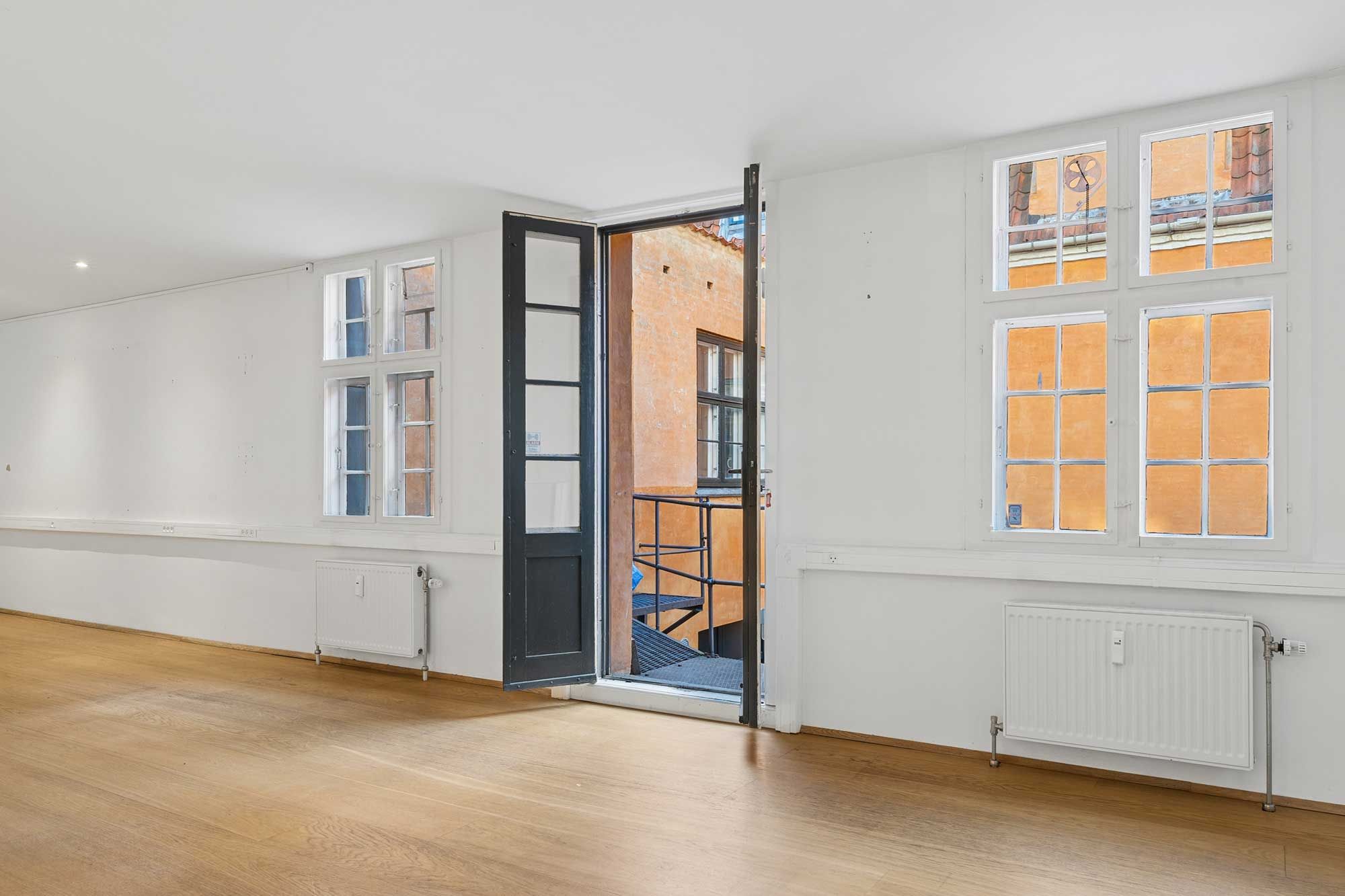 En tom, lyse, rummelig stue med højloft og trægulv. Der er tre vinduer og en dør, der er åben og viser til en trappe og et gårdrum med lysegulv