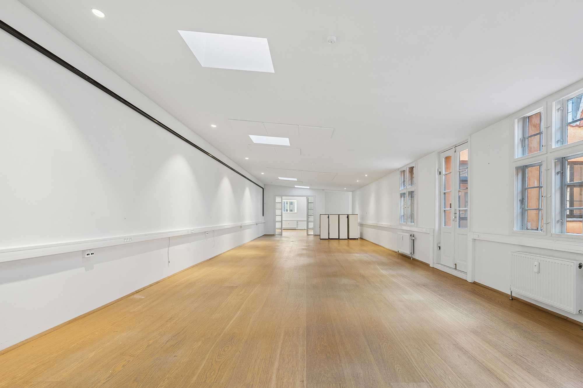 Stor kontorlokal i et hyggeligt gårdmiljø med lys, moderne indretning, lyse gulve, hvide vægge og store ruder. Lokalet har fuld loftshøjde og en luksuriøs følelse