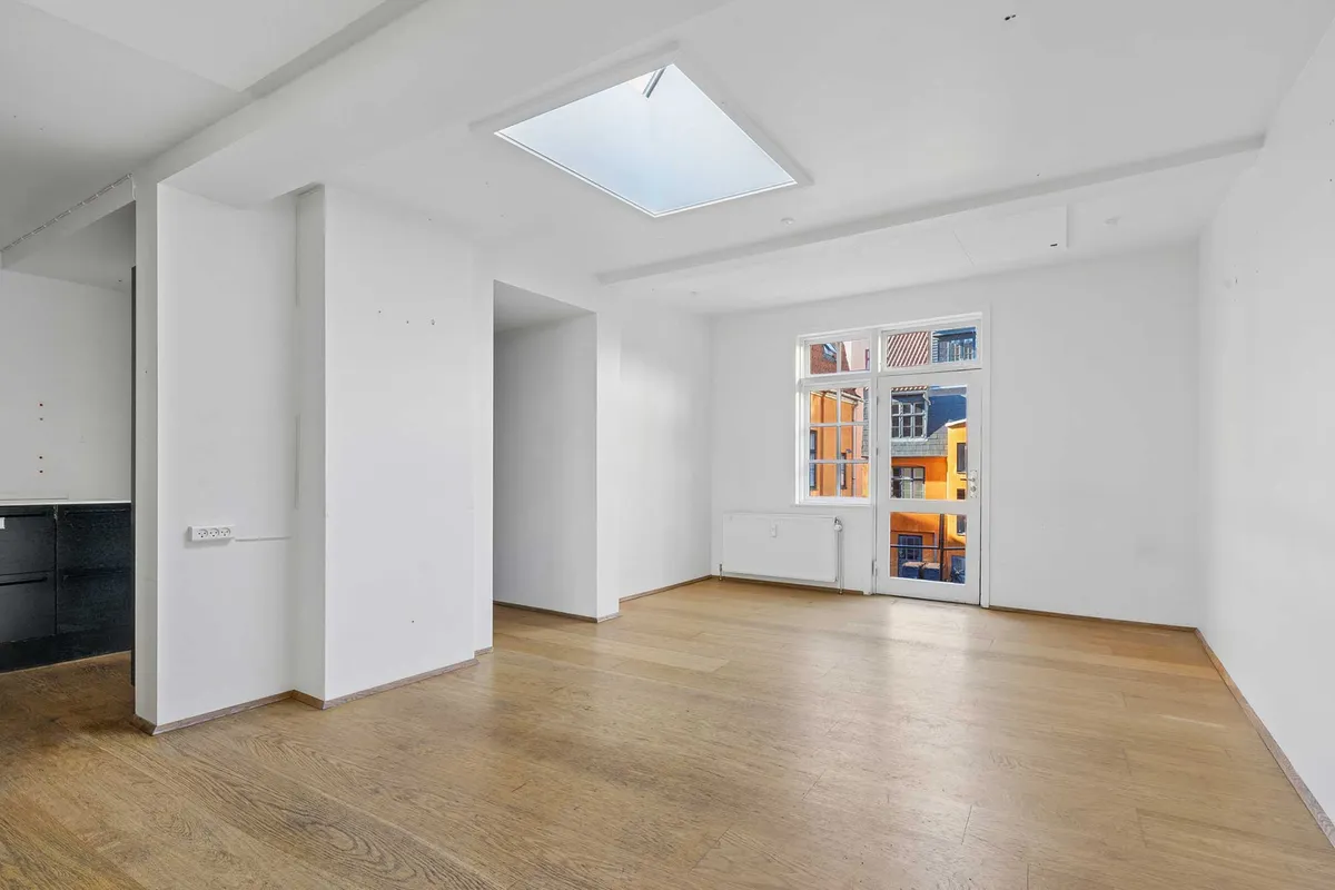 Et stort, lyse værelse med gulvtæppe, lyse gulvbelægning og store vinduer. Lystag og loftsvindue sørger for dagslys. Væggene er hvide og rummet er rent og ubenyttet