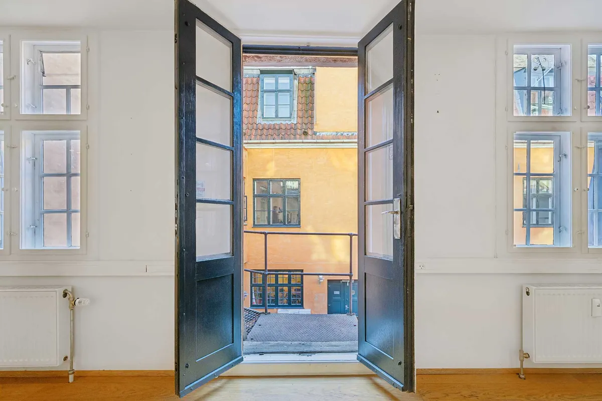 Et åbent kontordør med udsigt til en gård med gule vægge og rødt tag. Værelset er tomt med hvide vægge, gulve og radiatorer samt flere vinduer med udsigt til en gård.