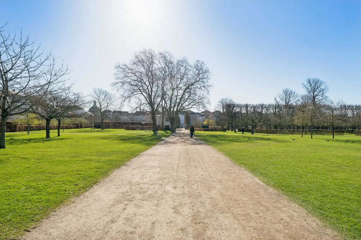 Grøn slette med stier og planter i solrigt vejr. Personer vandrer og en bygning i baggrunden. Træer med løv og græs samt spor af menneskelig aktivitet.