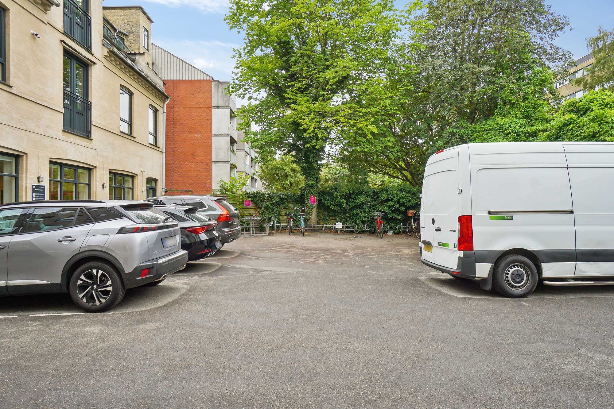Parkeringsplads med flere parkerede biler og en stor hvid varevogn i dagslys, omgivet af grønne planter og kontorbygninger