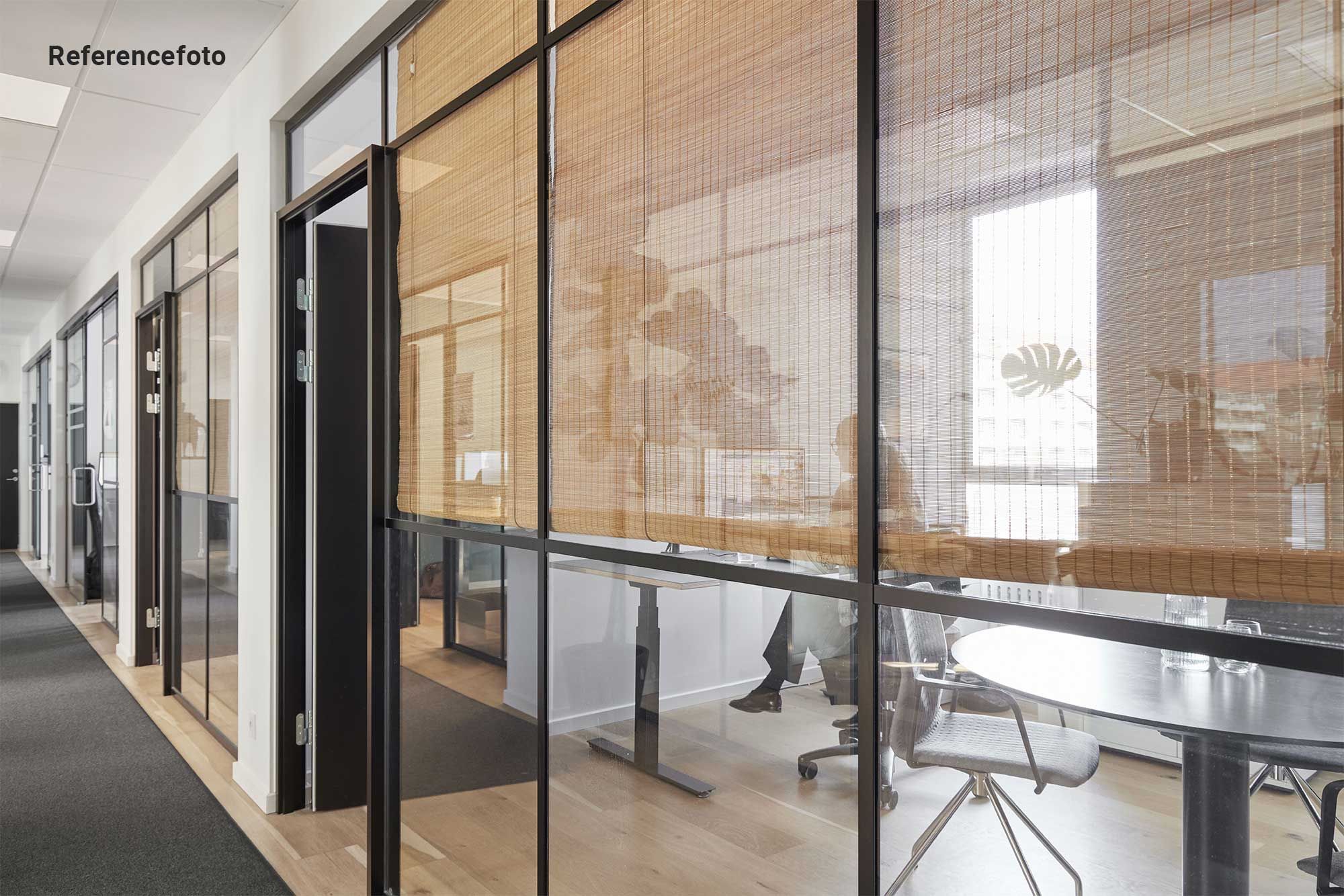 Kontor i tidligere cigarfabrik med trægulv og glasvægge. Glasruder og ruller med bambusmønstre. Bord med glas og stole. Indvendigt plante og vinduer med udsigt.