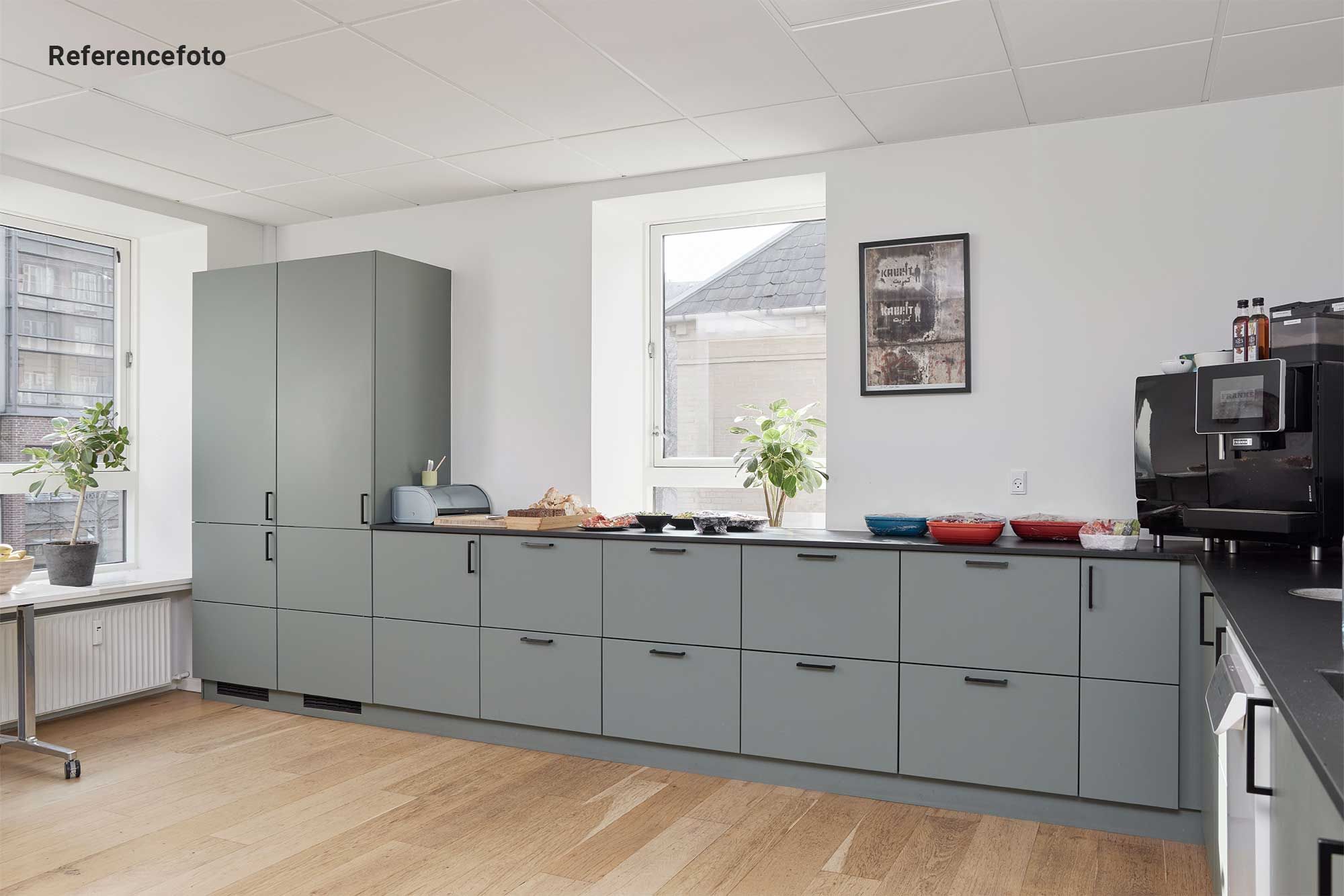 Moderne kontor med hvidt loft, hårdt trægulv og grå kabinetter med håndtag. På en bordplade er der en mikrobølgeovn, en kopimaskine og grønne planter.