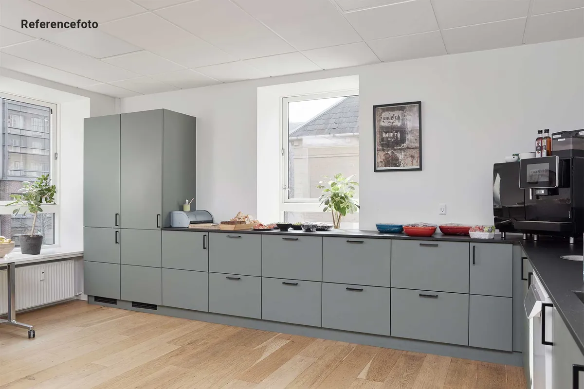 Moderne kontor med hvidt loft, hårdt trægulv og grå kabinetter med håndtag. På en bordplade er der en mikrobølgeovn, en kopimaskine og grønne planter.