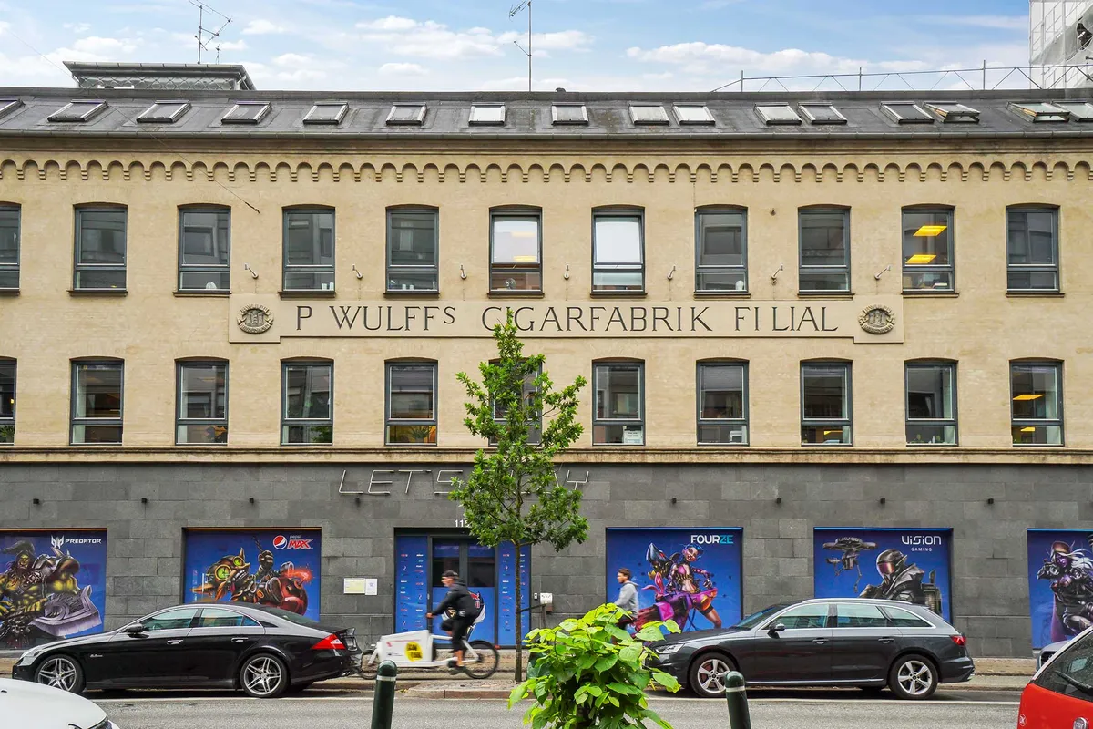 To biler parkeret på gaden foran P. Wulffs Cigarfabrik. En mand kører på en cykel med en kuffert og en kvinde går foran ham. Bygningen med flere vinduer har flere reklameplakater