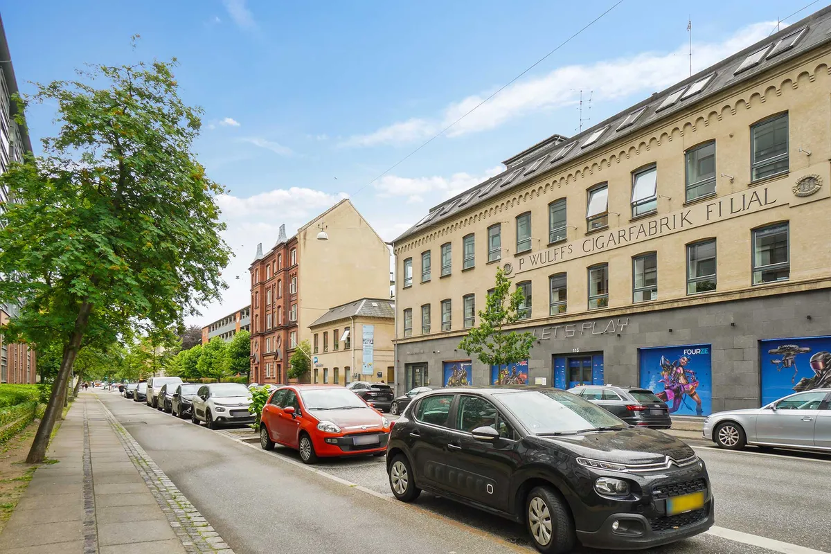 Parkerede biler står på gaden foran en bygning med facaden P. Wulffs Cigarfabrik Filial. Bygningen er i beton og gult murværk med vinduer og solceller