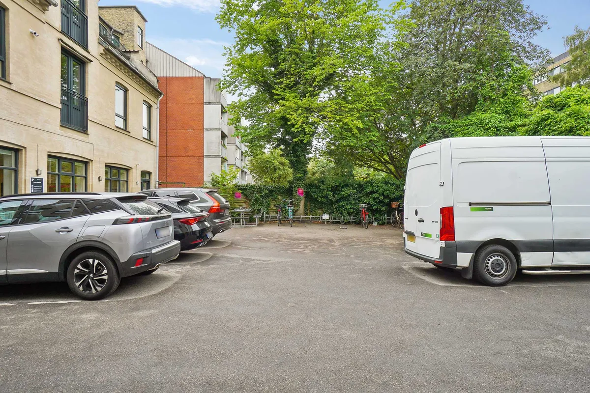 Parkerede biler i en grøn zone med en stor hvid lastbil i baggrunden og to cykler i en cykelholder