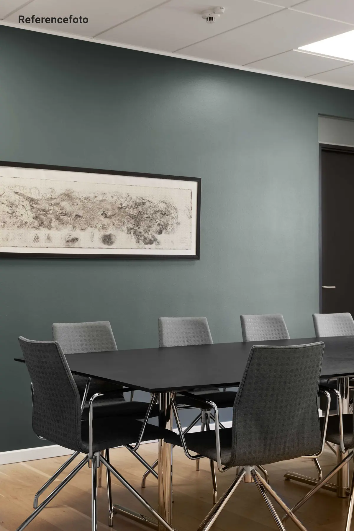 Kontormøbler med høje stolben og tynd metalramme. Bord med høje ben og slank kant. Væg i lyseblå og ramme om kunstværk. Hvid loftsbeklædning og gul trægulv.