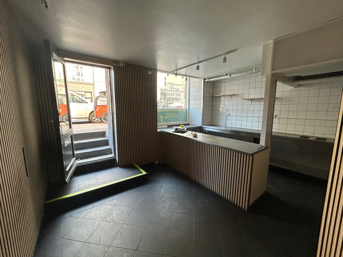 Take-away-lokale med lyse blyegrønne vægge og lysegrå møbler. Butik med åben dør og trin op til gulvet. Køkken med hvide kakelvægge og lysstribe i loftet