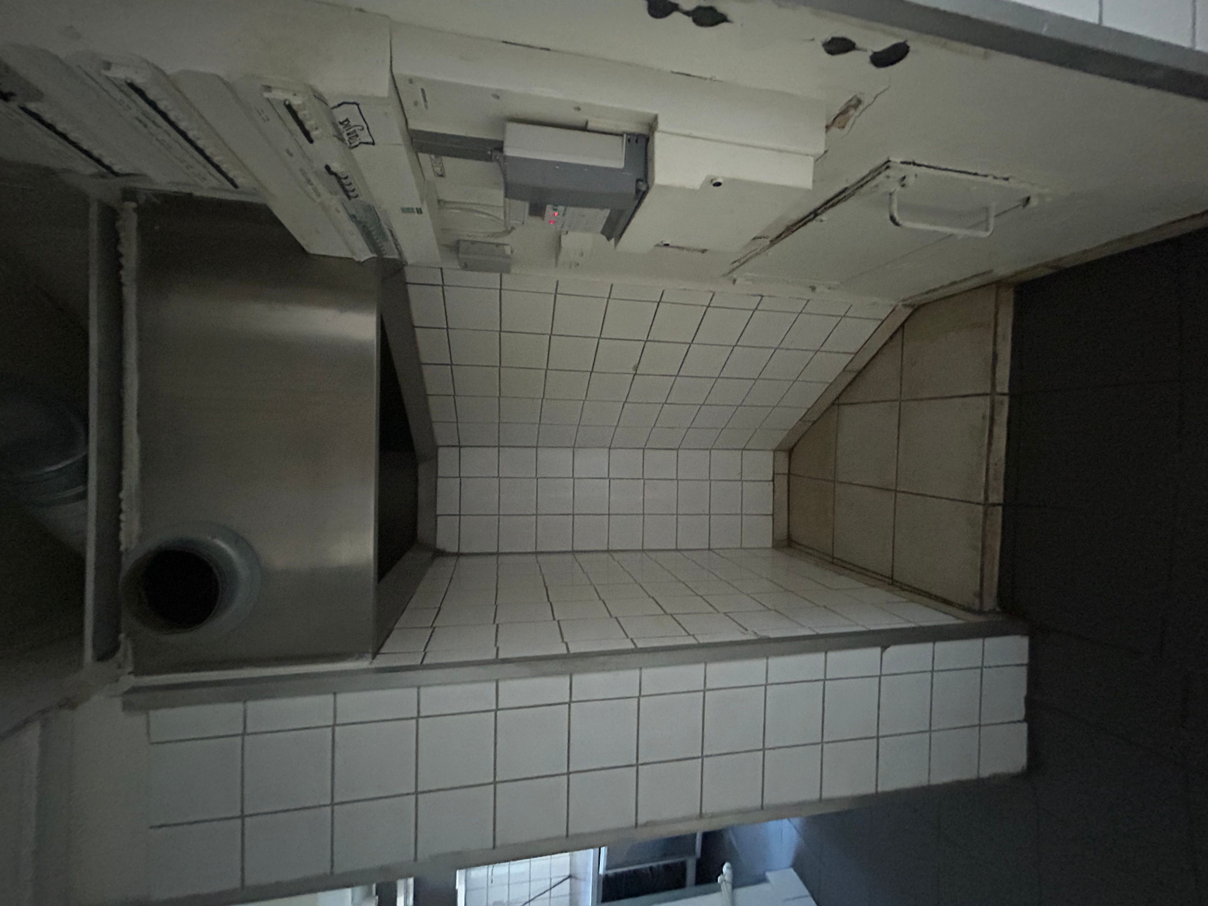 Hjørne af køkken med udsugningsventilator og hvidt fliser og gulv. Rum med mørkt loft og forskellige elektriske apparater. Hjørne med skrånende væg og højdeforskel