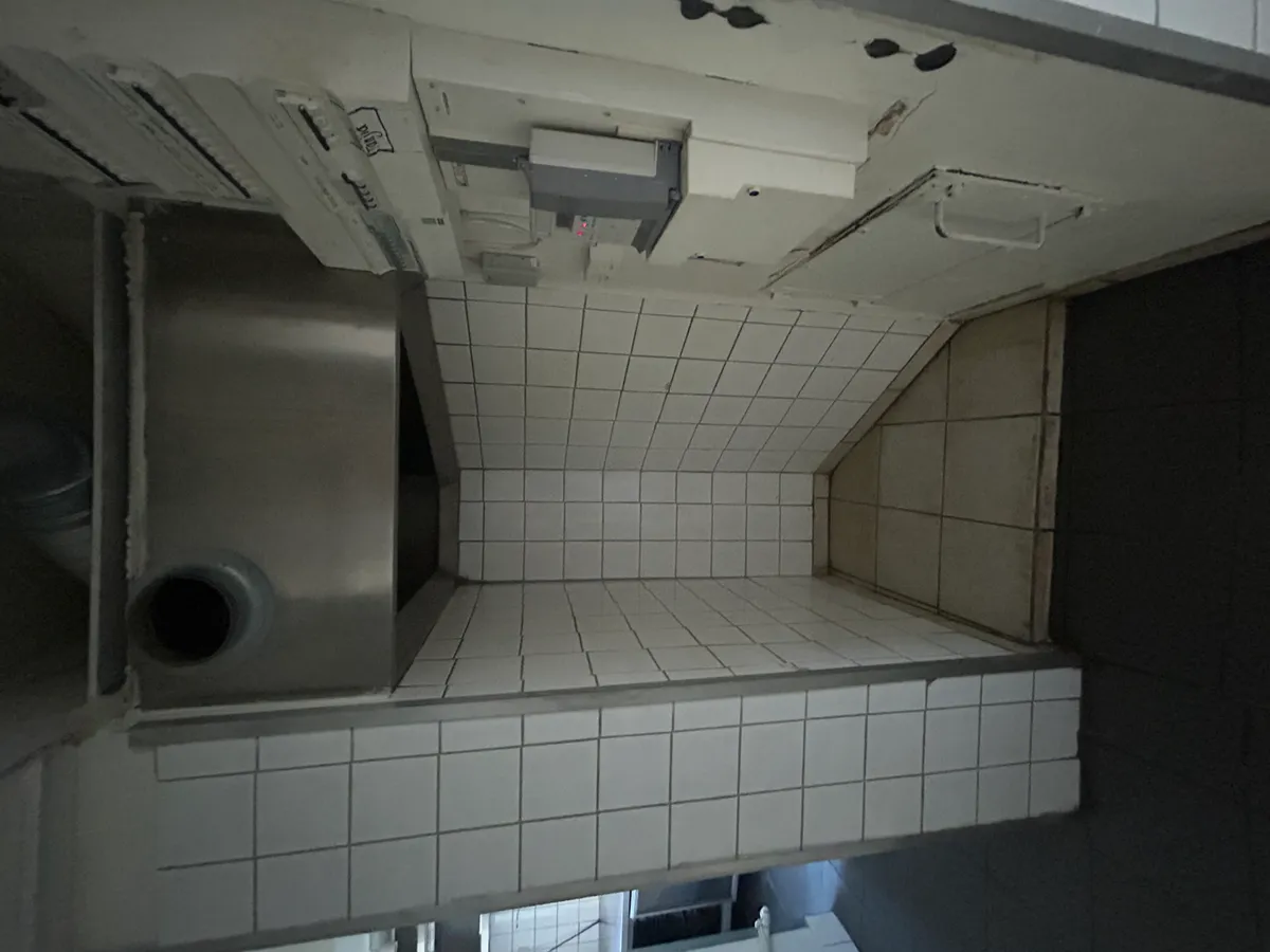 Hjørne af køkken med udsugningsventilator og hvidt fliser og gulv. Rum med mørkt loft og forskellige elektriske apparater. Hjørne med skrånende væg og højdeforskel