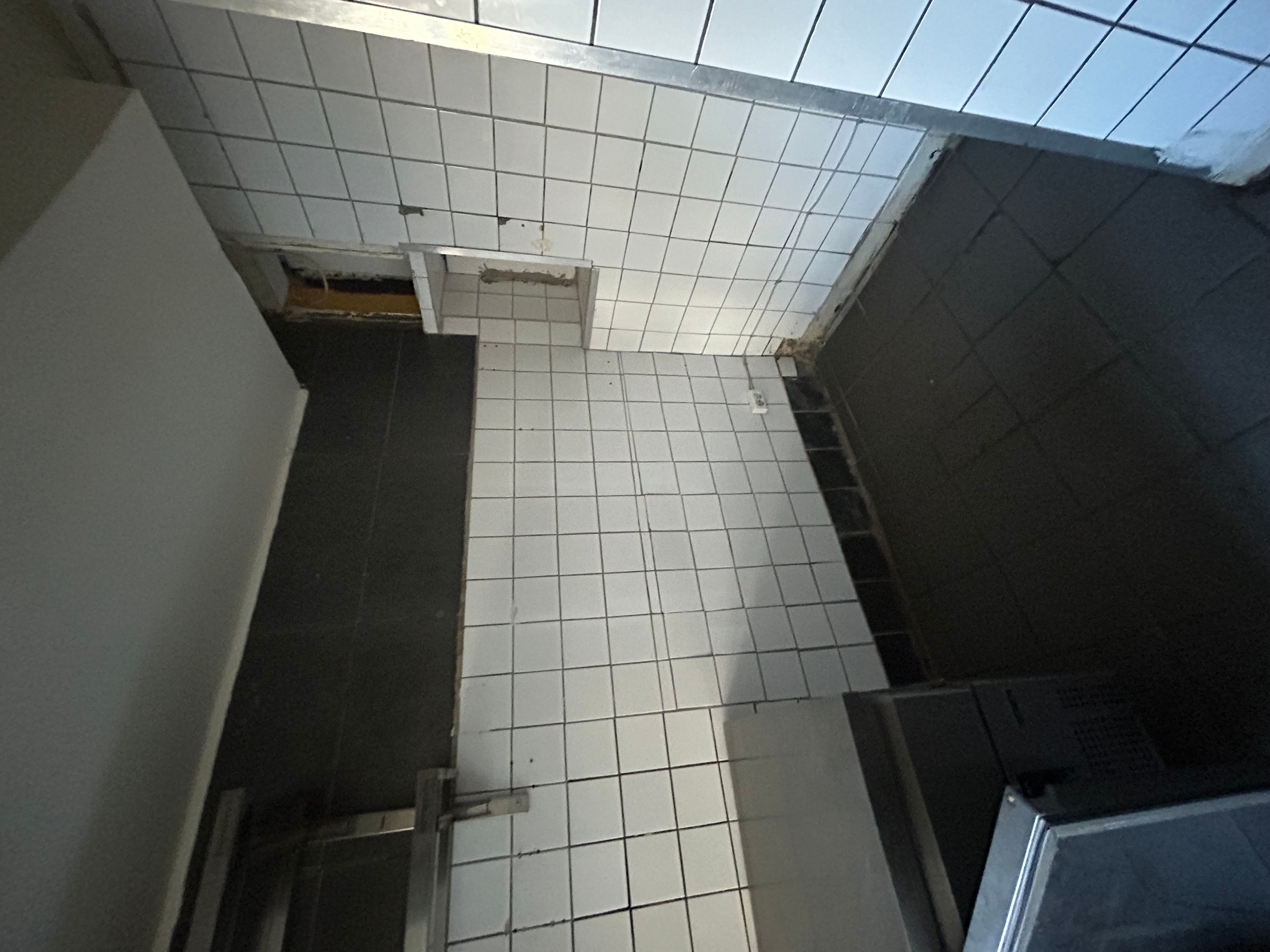 Et hjørne med hvide og sorte fliser og en hvid væg, der ender i et mørkt gulv. Lokalet er delvist renoveret med åbninger og udstyr i gangen.