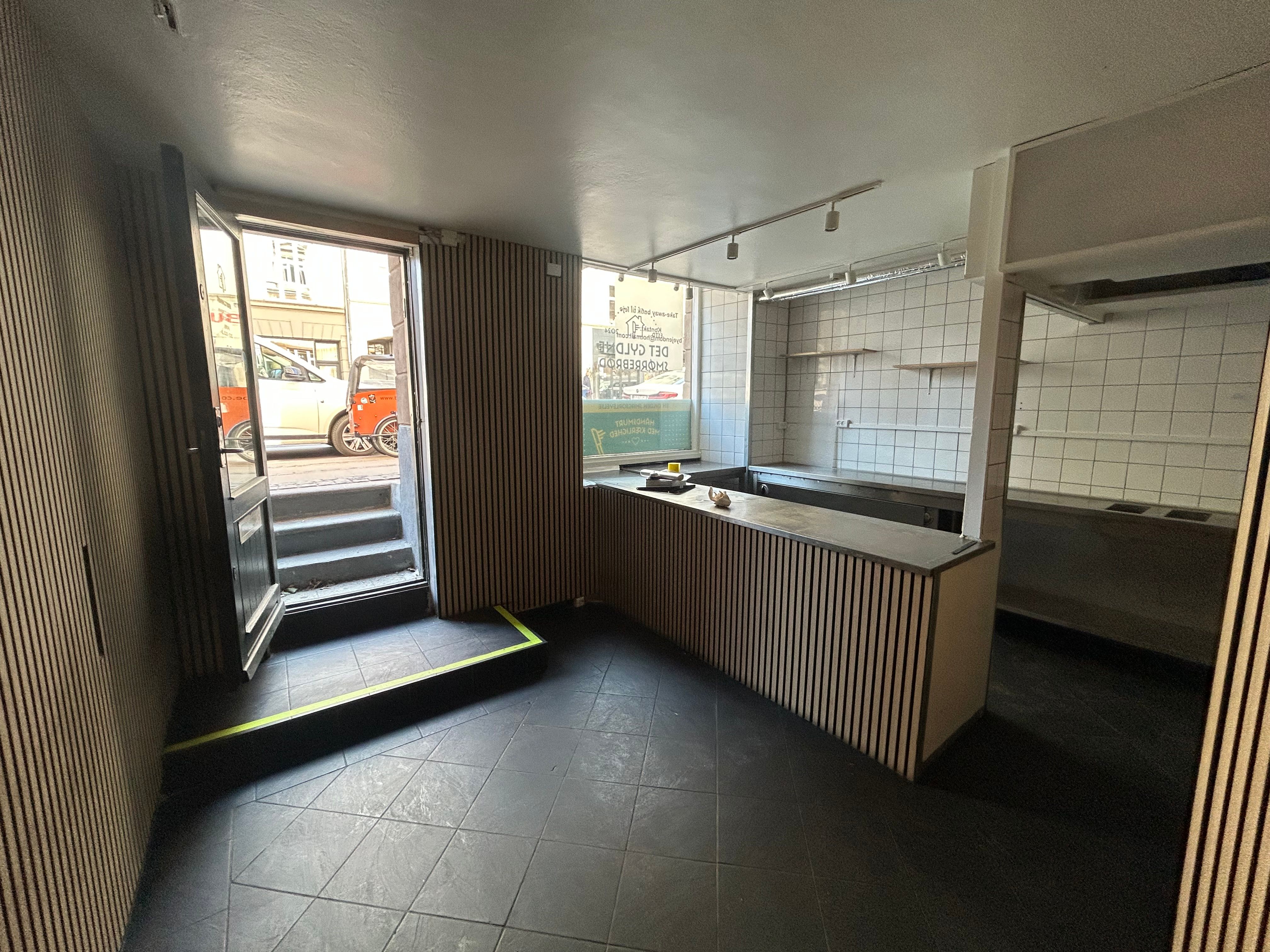 Take away lokal til leje med udsigt til gade med kørende biler og trin, åben dør og glasruder og møbler i rummet