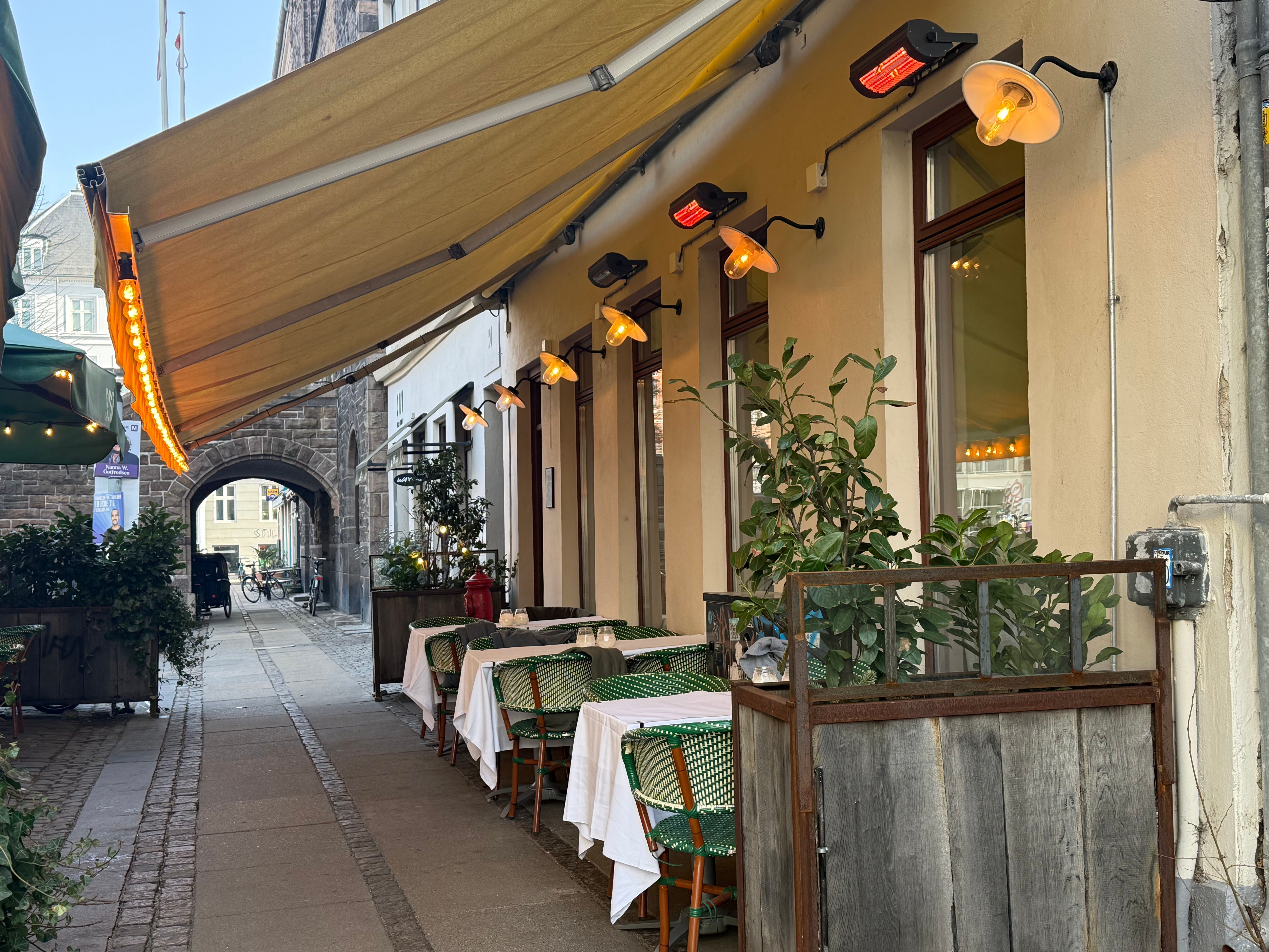 Lejemål for restaurant på Vesterbrogade 51 med solbeskyttelse, lysekroner og grønne planter langs gaden