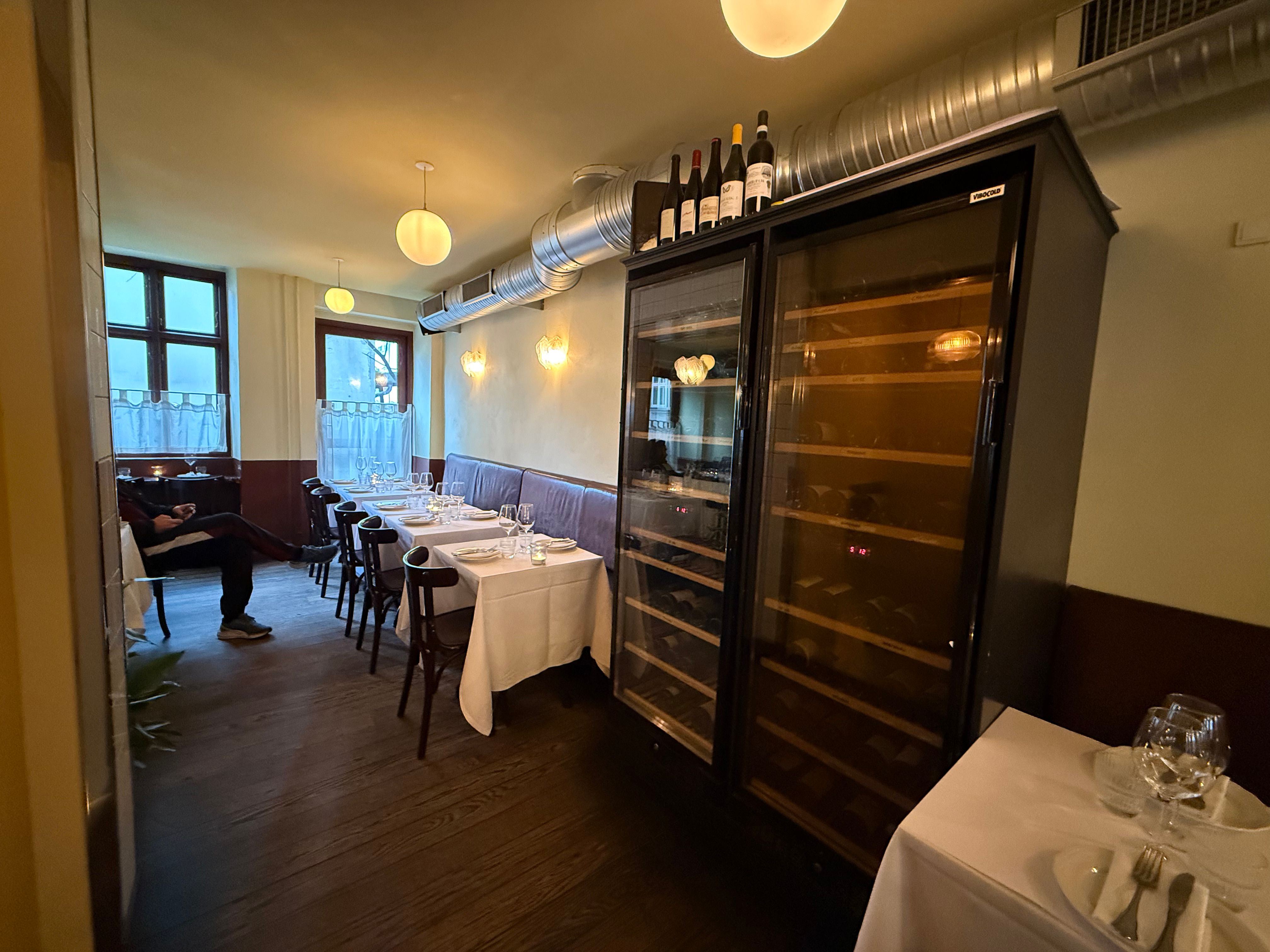 En elegant tom restaurant med bord og stol, glasvinduer, lysekroner og et større vinskabssystem med vinflasker på toppen