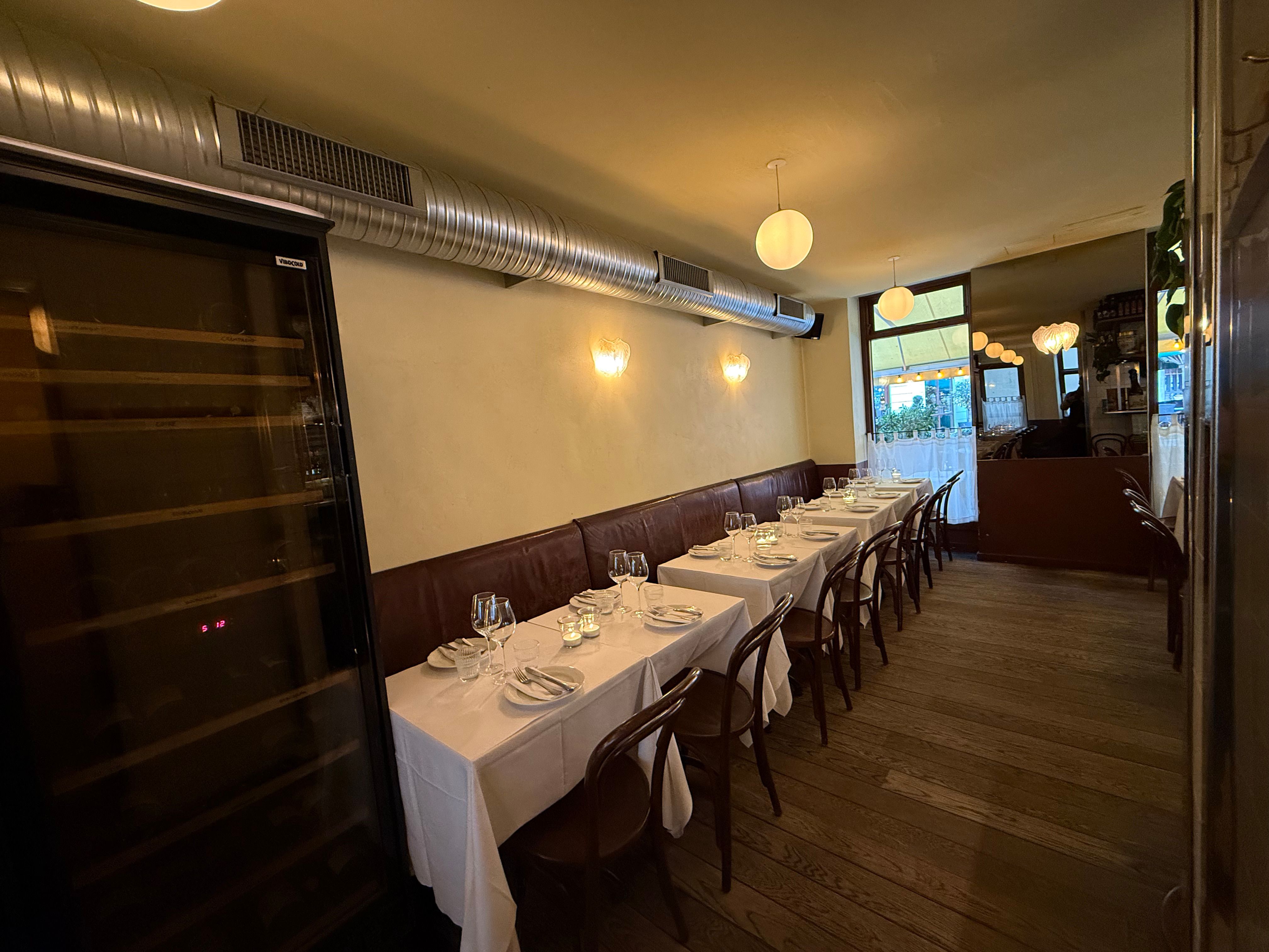 En elegant restaurant med hvide borddæksler og lyse gulvvægge, udstyret med lysekroner og vinduer med udsigt til gaden