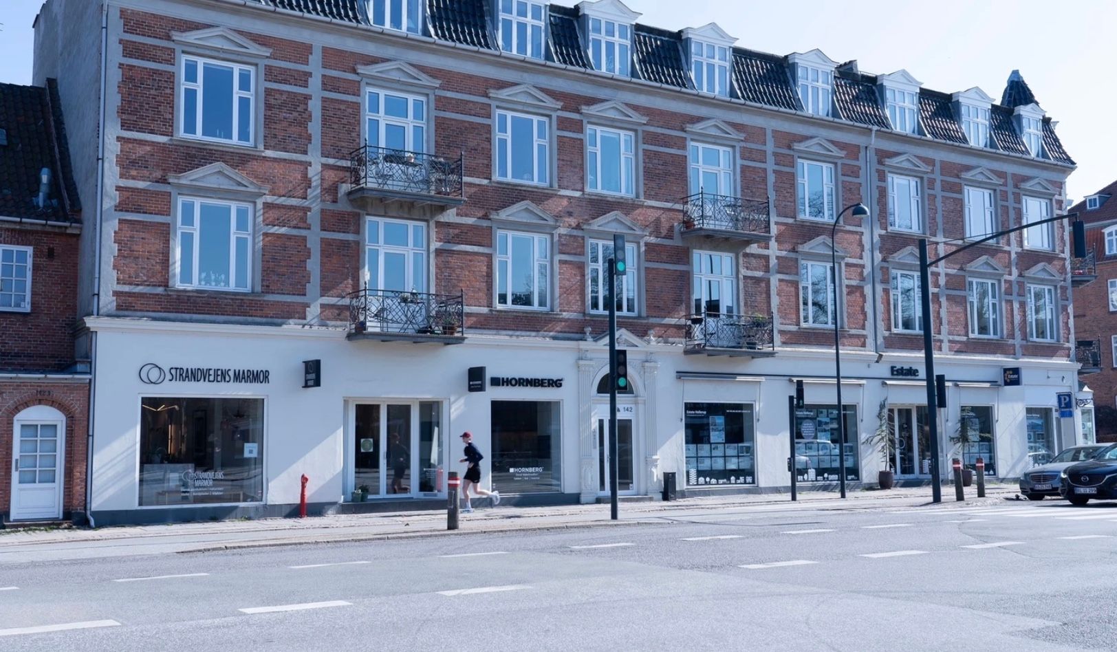 To butikker med skilte Strandvejens Marmor og Hornberg på Strandvejen i Hellerup. En person løber udenfor, og der er biler parkeret. To gadelampestænger med trafiklys står på hjørnet.