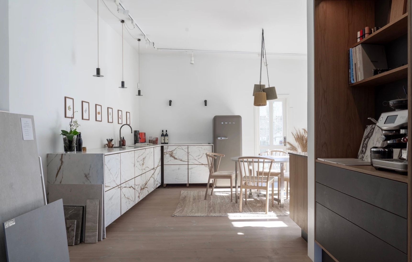 Foto af moderne og minimalistisk køkken med marmor køkkenbord, hvidt køkkenskab og loftslys. Smukke træstole, bord og gulvtæppe foran og køleskab med lysekroner og små kunstværker på væggen