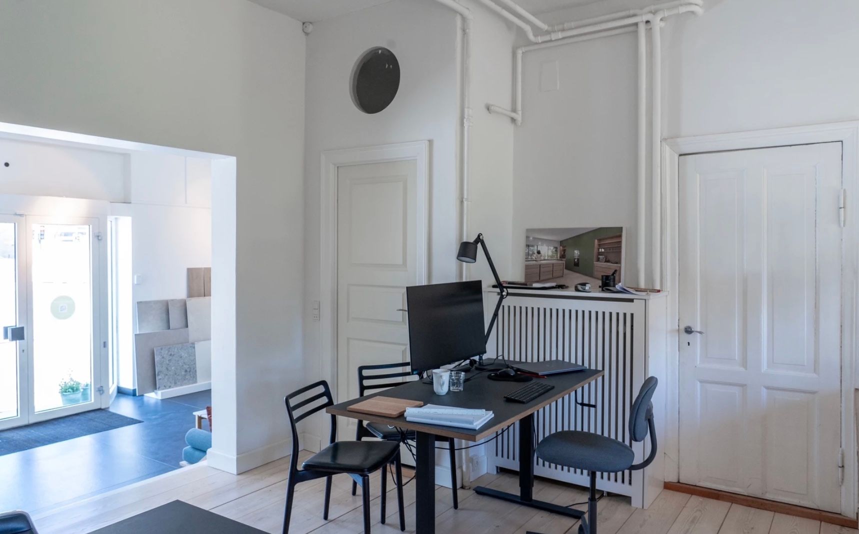 Foto af en møbleret kontorrum med en hvid væg, hvor der er et bord med en computer og en lampe