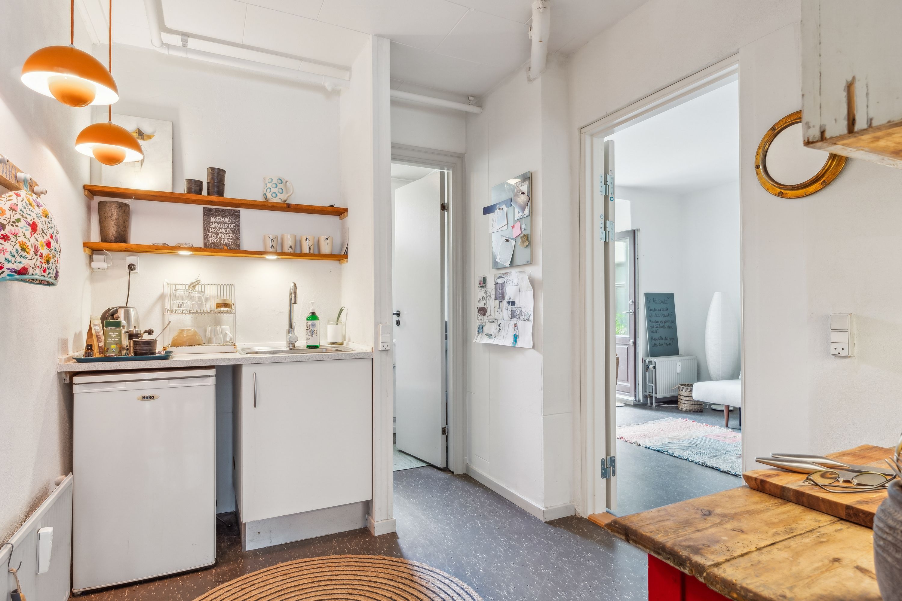 Hvid indretning med køkken med lysekroner og gulvtæppe, skraldespand og køleskab, åben dør og lyse gulve samt køkkenredskaber