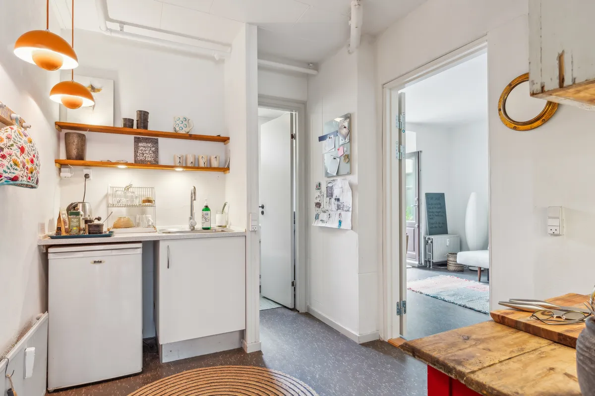 Hvid indretning med køkken med lysekroner og gulvtæppe, skraldespand og køleskab, åben dør og lyse gulve samt køkkenredskaber