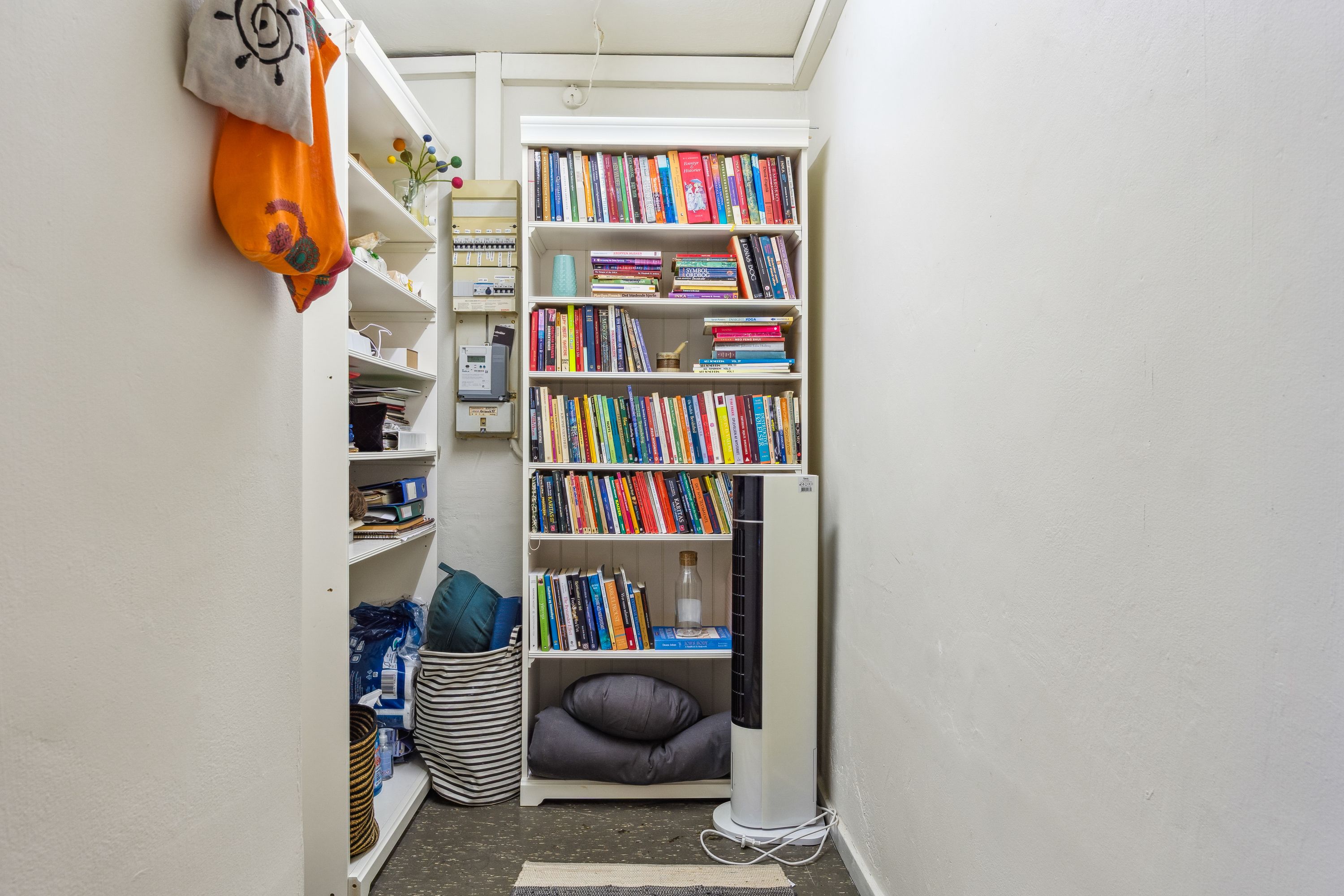 Lille bibliotek med mange bøger, hvor der er liggende tæpper og pude på gulvet og en fan. Væggene er hvide og der er en række hylder med bøger, tæpper, kugle, tøj og papirkurv