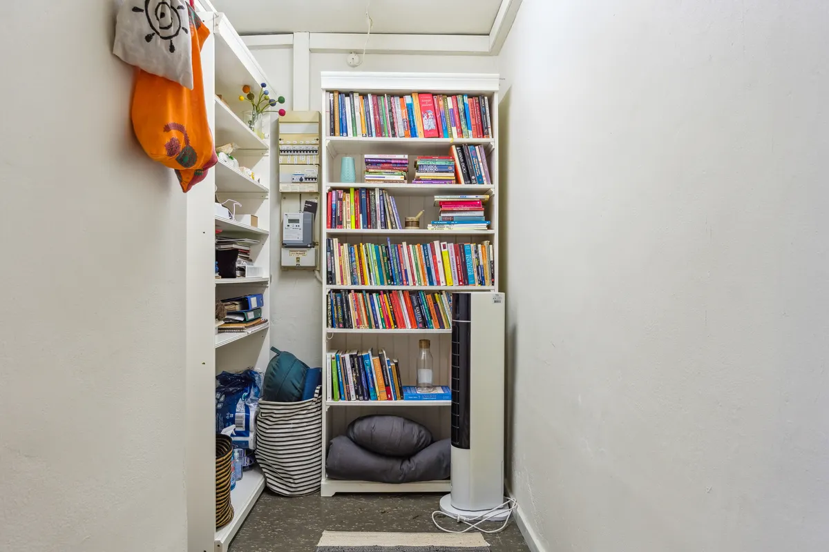 Lille bibliotek med mange bøger, hvor der er liggende tæpper og pude på gulvet og en fan. Væggene er hvide og der er en række hylder med bøger, tæpper, kugle, tøj og papirkurv