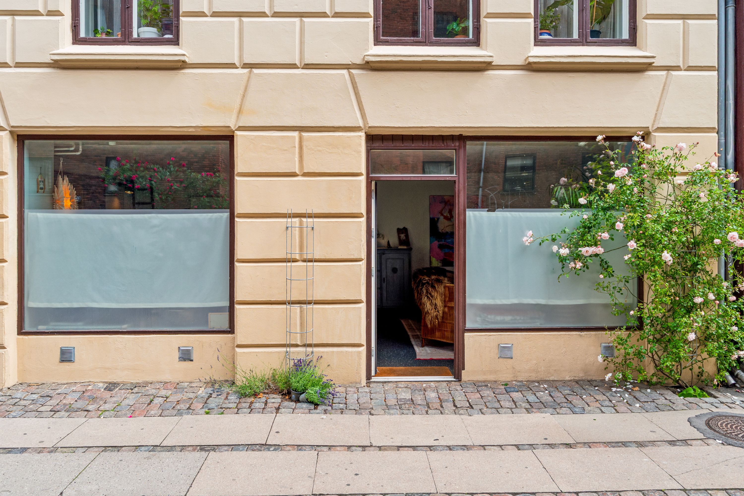 Butik på Korsgade 10 i København med køkkenbord og blomster i vinduet og en større urtepotte på gaden. Planter og buske på siden af bygningen og en åben dør