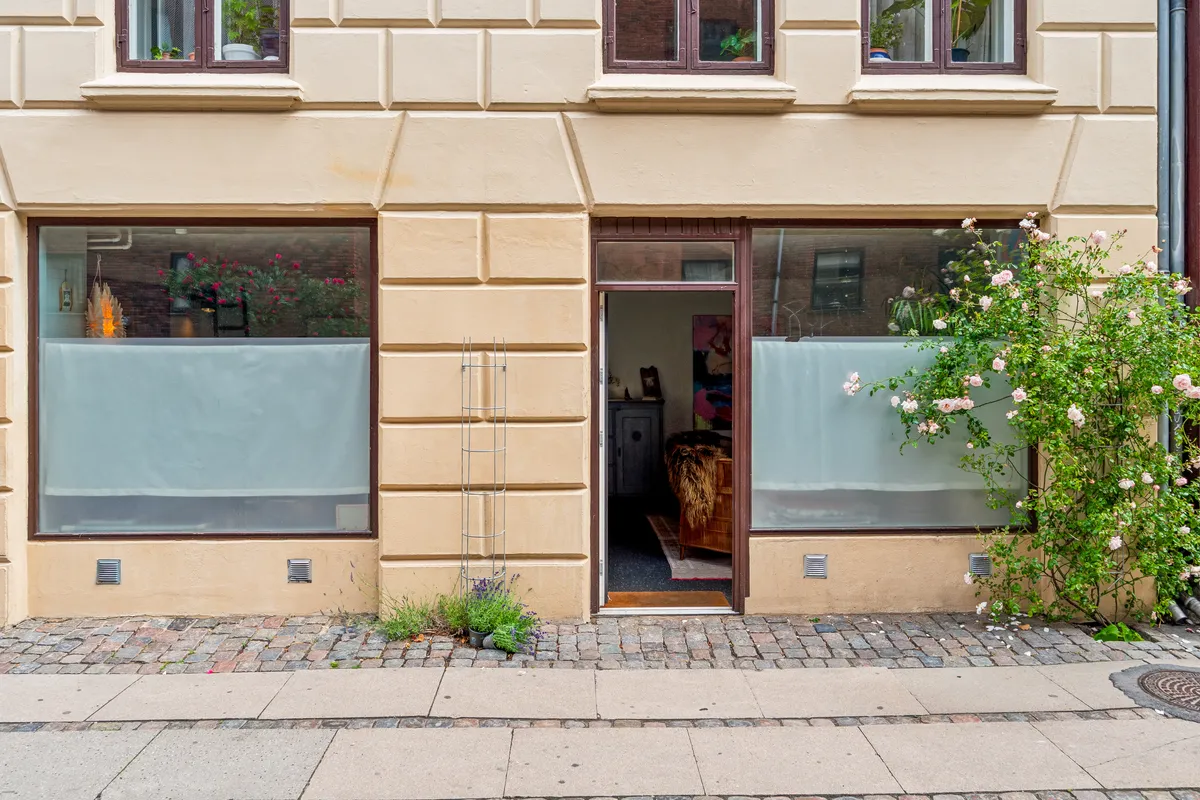 Butik på Korsgade 10 i København med køkkenbord og blomster i vinduet og en større urtepotte på gaden. Planter og buske på siden af bygningen og en åben dør